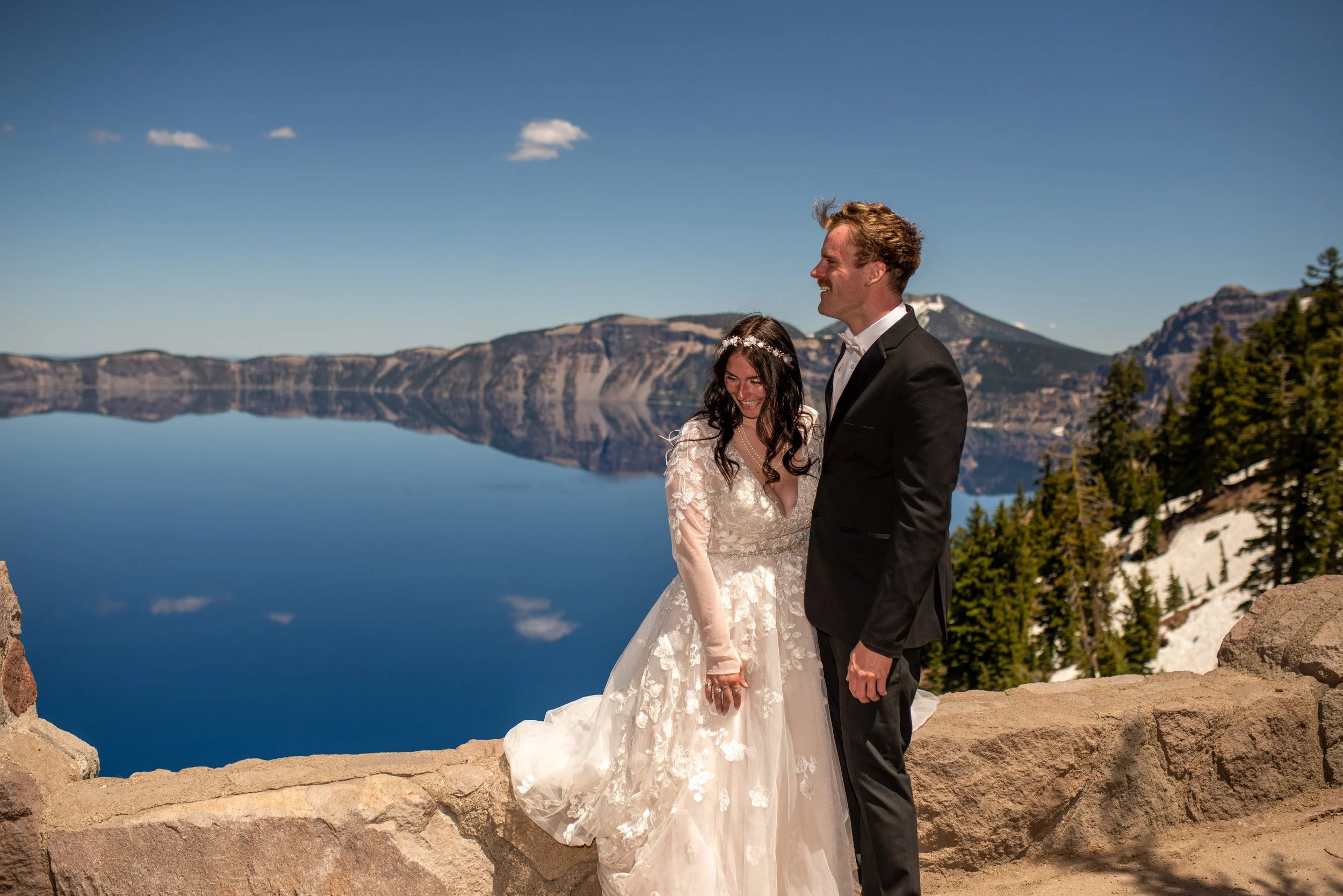 A bride and groom standing outdoors near a rocky ledge with a lake and mountain landscape in the background, smiling and holding hands during their wedding.