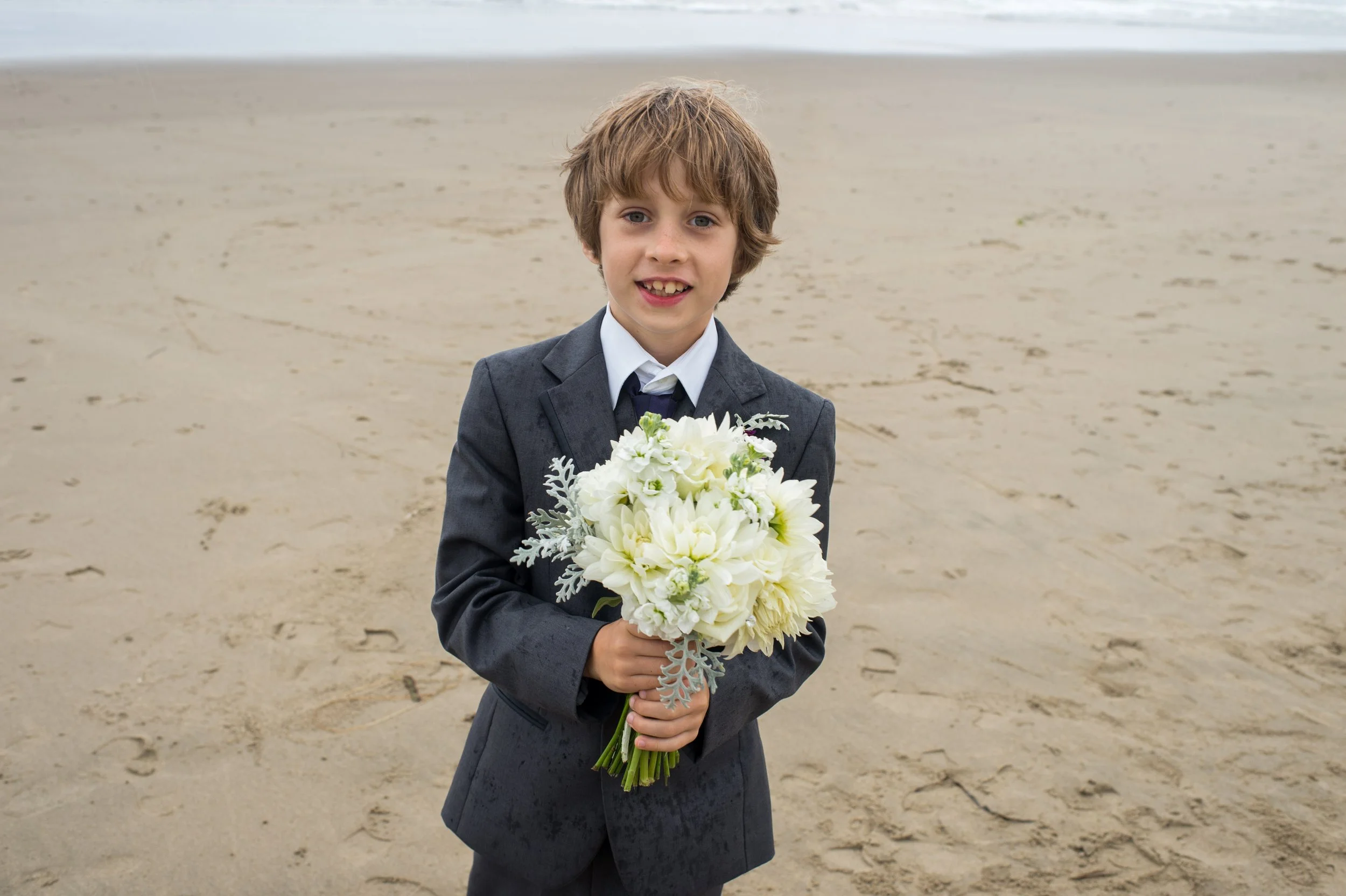 A young boy dressed in a gray suit holding a bouquet of white flowers on a sandy beach.