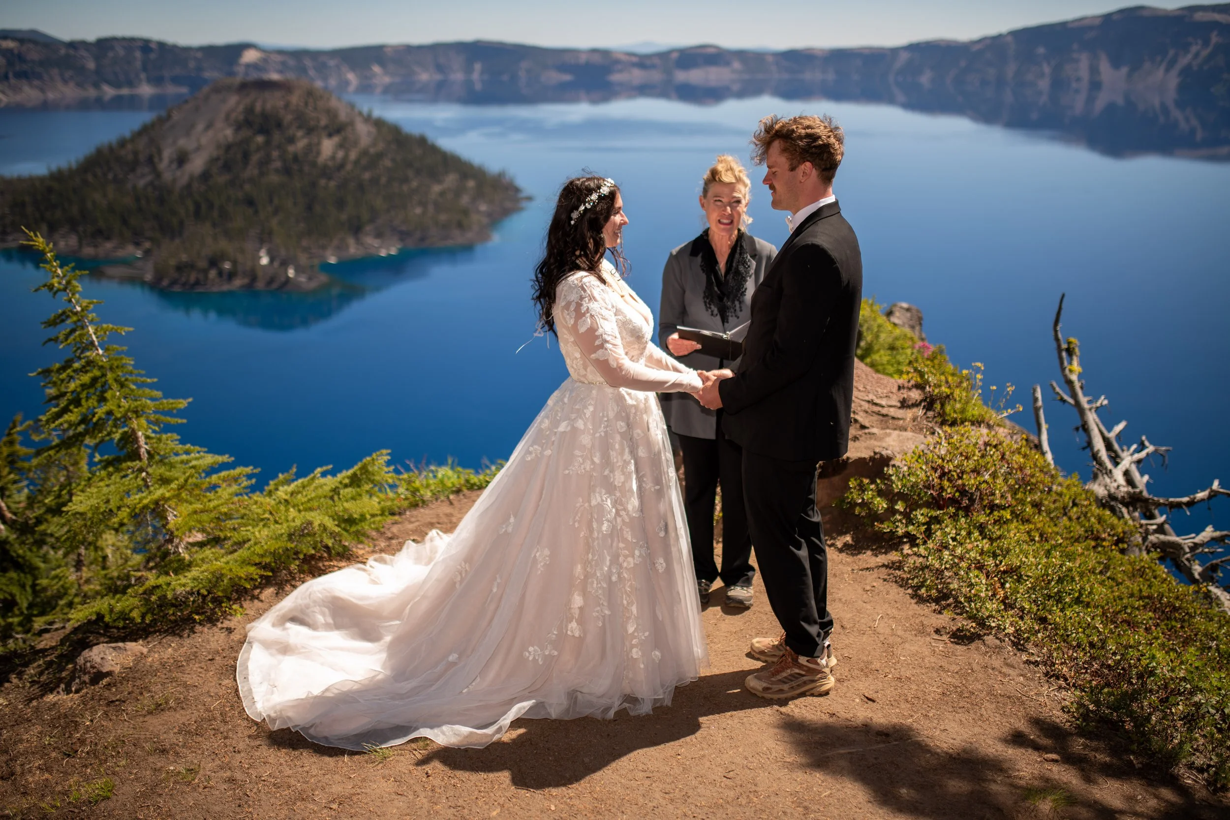 A wedding ceremony is taking place outdoors on a cliff overlooking a lake with mountains in the background. The bride and groom are facing each other, holding hands, with a officiant standing between them. The bride wears a white wedding dress with l