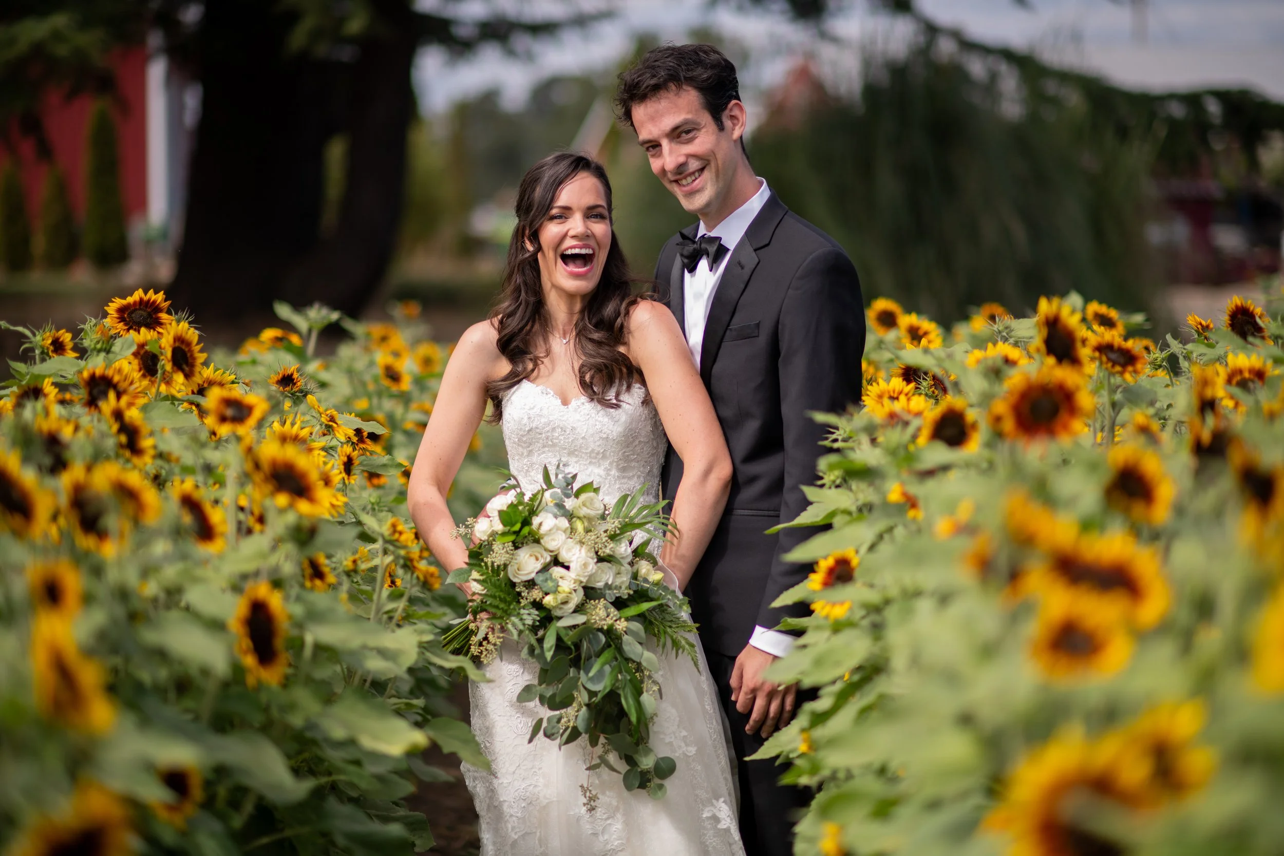 A newlywed couple standing in a sunflower field, with the bride in a white wedding dress holding a bouquet of white roses and greenery, and the groom in a black tuxedo with a bow tie, smiling and posing together.