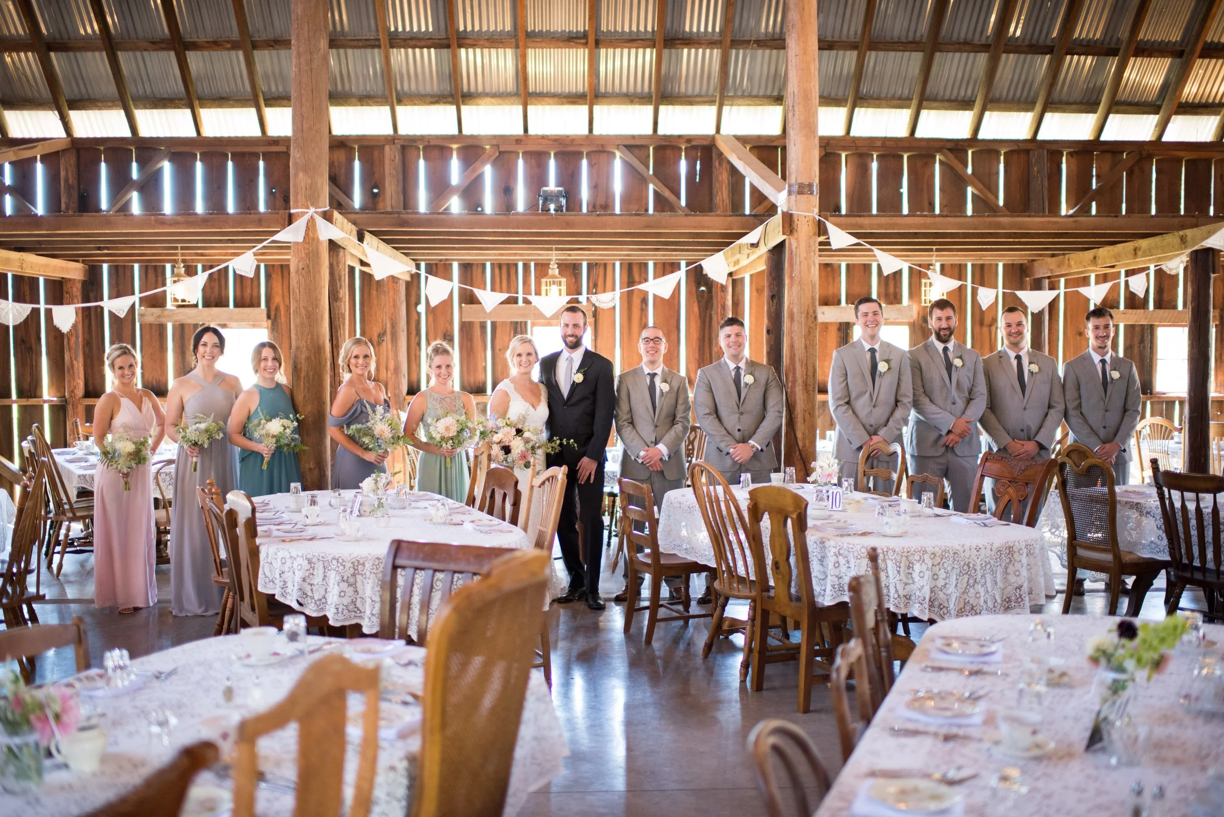 People gathered in a rustic barn decorated for a wedding, standing in a line, dressed in formal attire, with floral bouquets in hand, surrounded by decorated tables and string lights.