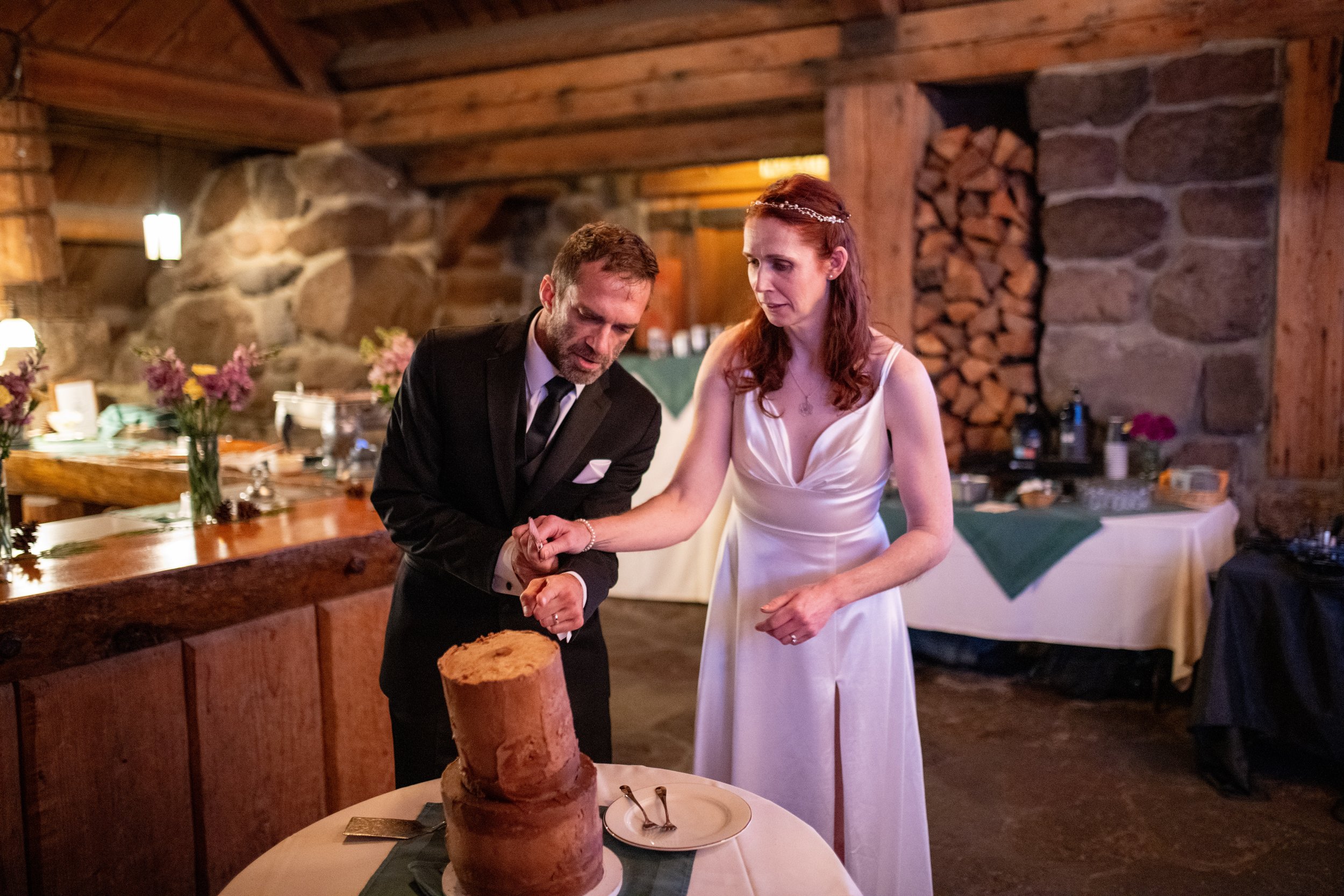 A groom in a black tuxedo and a bride in a white wedding dress are cutting a cake together at a wedding reception in a rustic venue with wooden beams and stone walls, decorated with flowers.