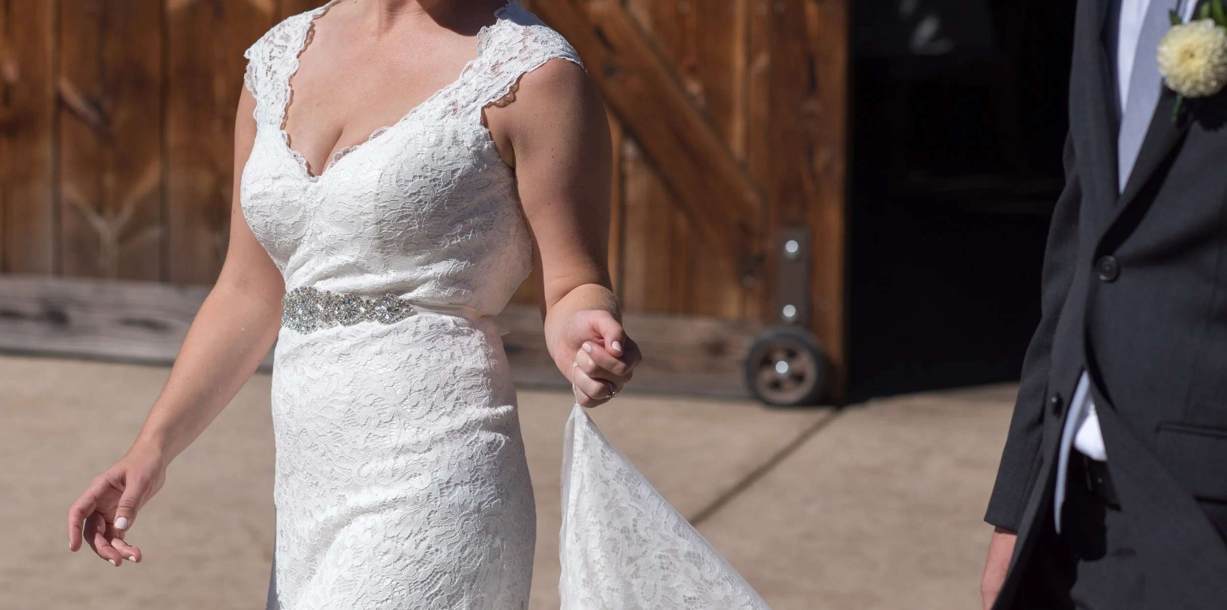 A bride in a white lace wedding gown with a jeweled waistband, holding her dress with one hand, standing outside next to a groom in a black tuxedo with a white boutonniere, on a sunny day with a wooden barn in the background.