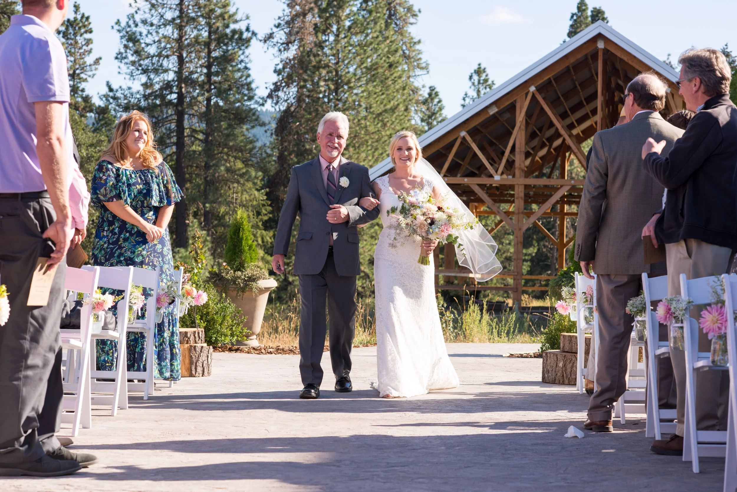 A bride walking down the aisle with an older man, likely her father, during an outdoor wedding ceremony in a forested area under a partly cloudy sky, with guests on either side.