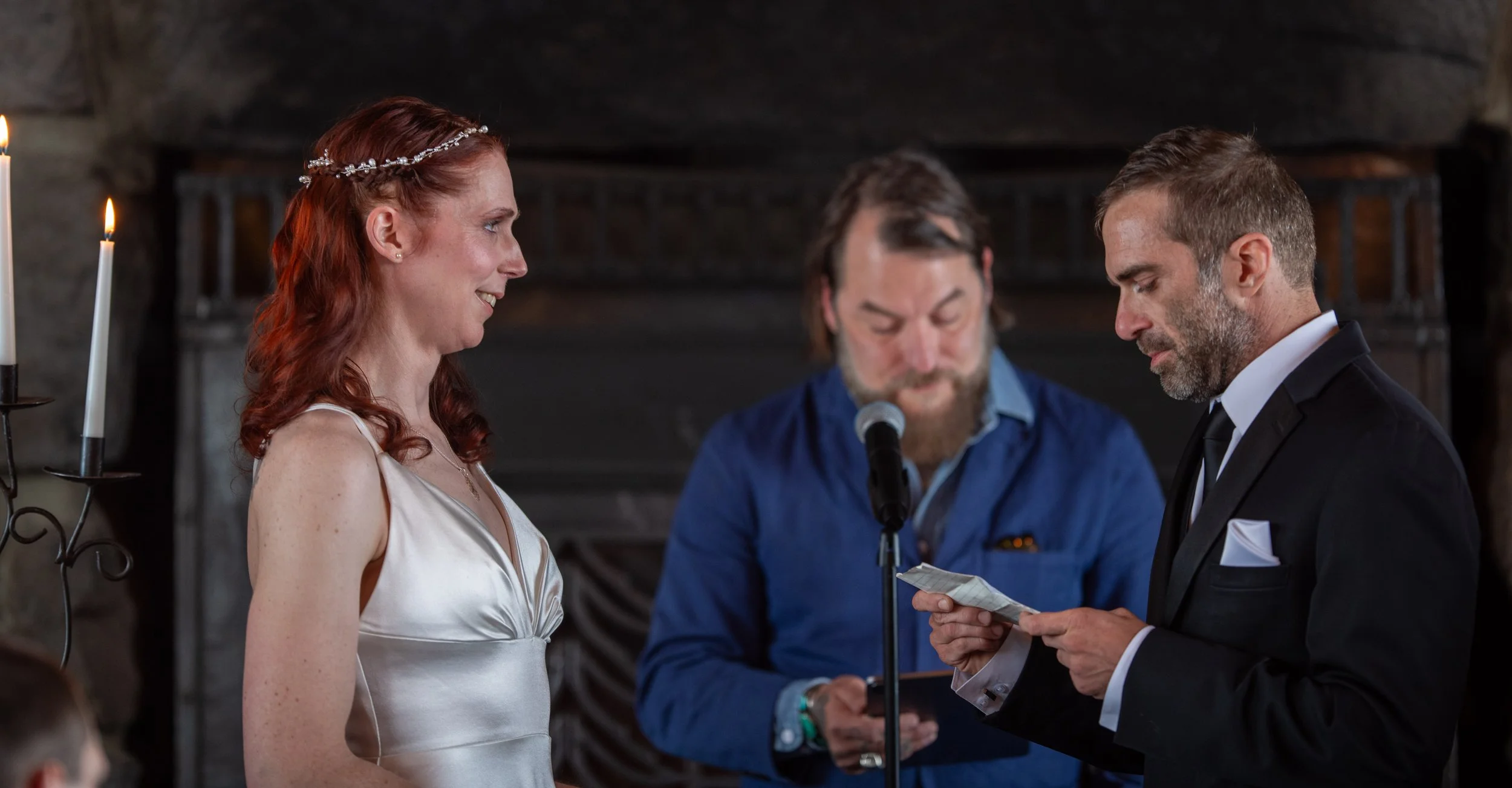 A wedding ceremony with a bride and groom exchanging vows, with an officiant reading from paper, inside a dimly lit room with candles.