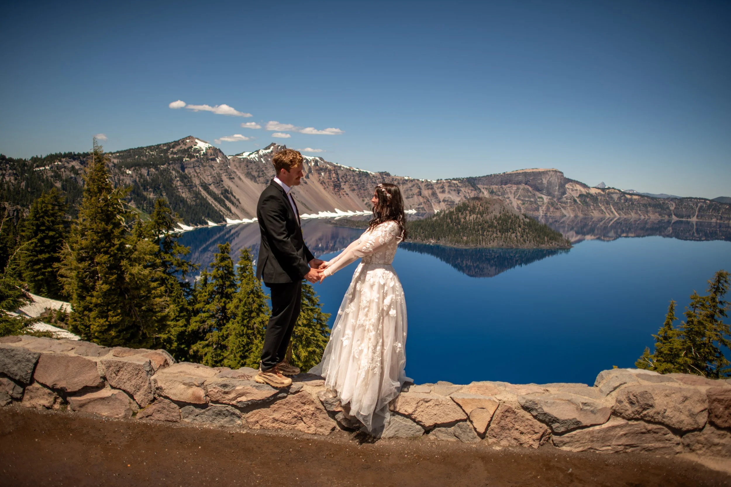 A couple in wedding attire holding hands and facing each other by a mountain lake with snow-capped peaks in the background.