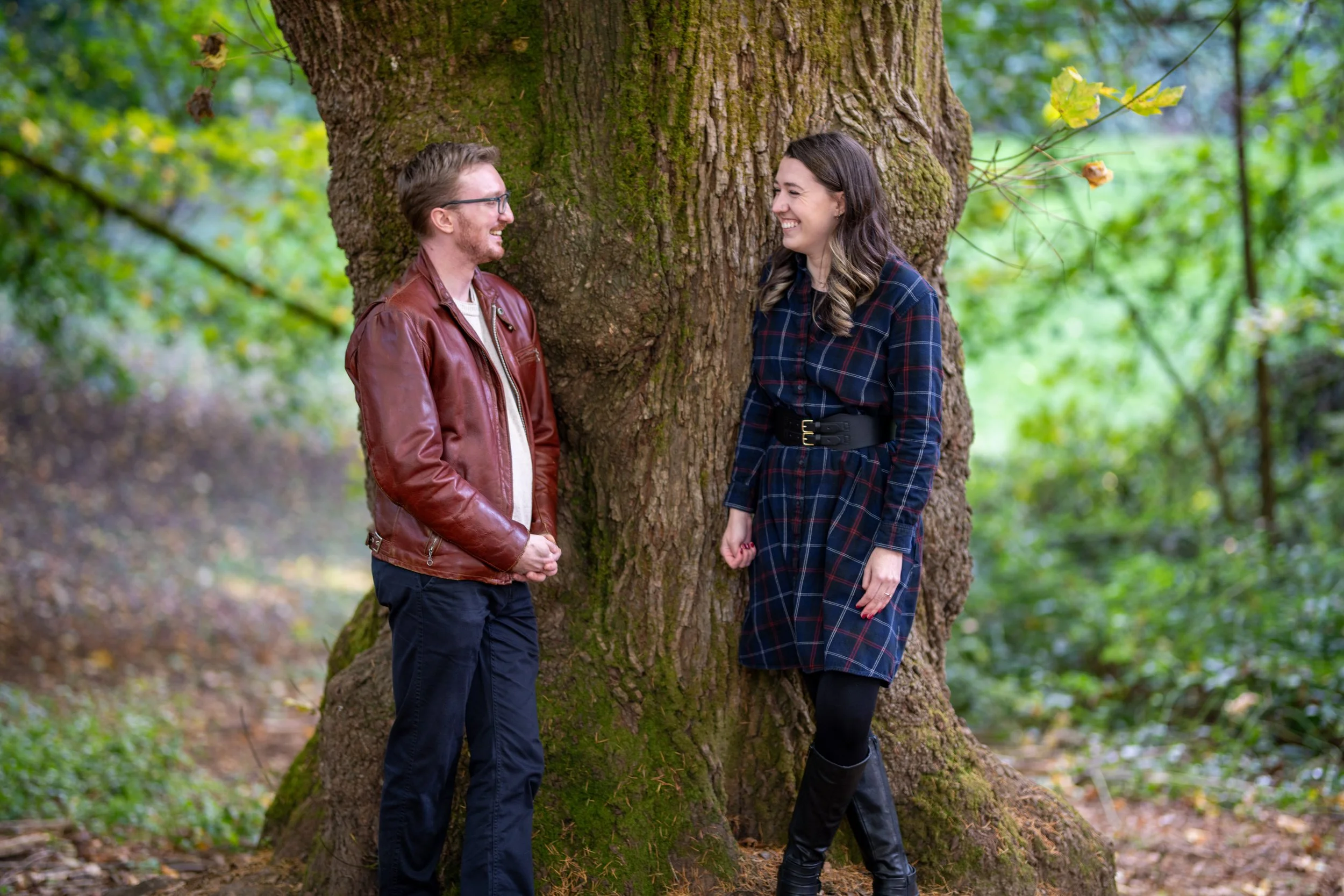 A couple standing by a large tree in a forest, smiling at each other, with green foliage in the background.