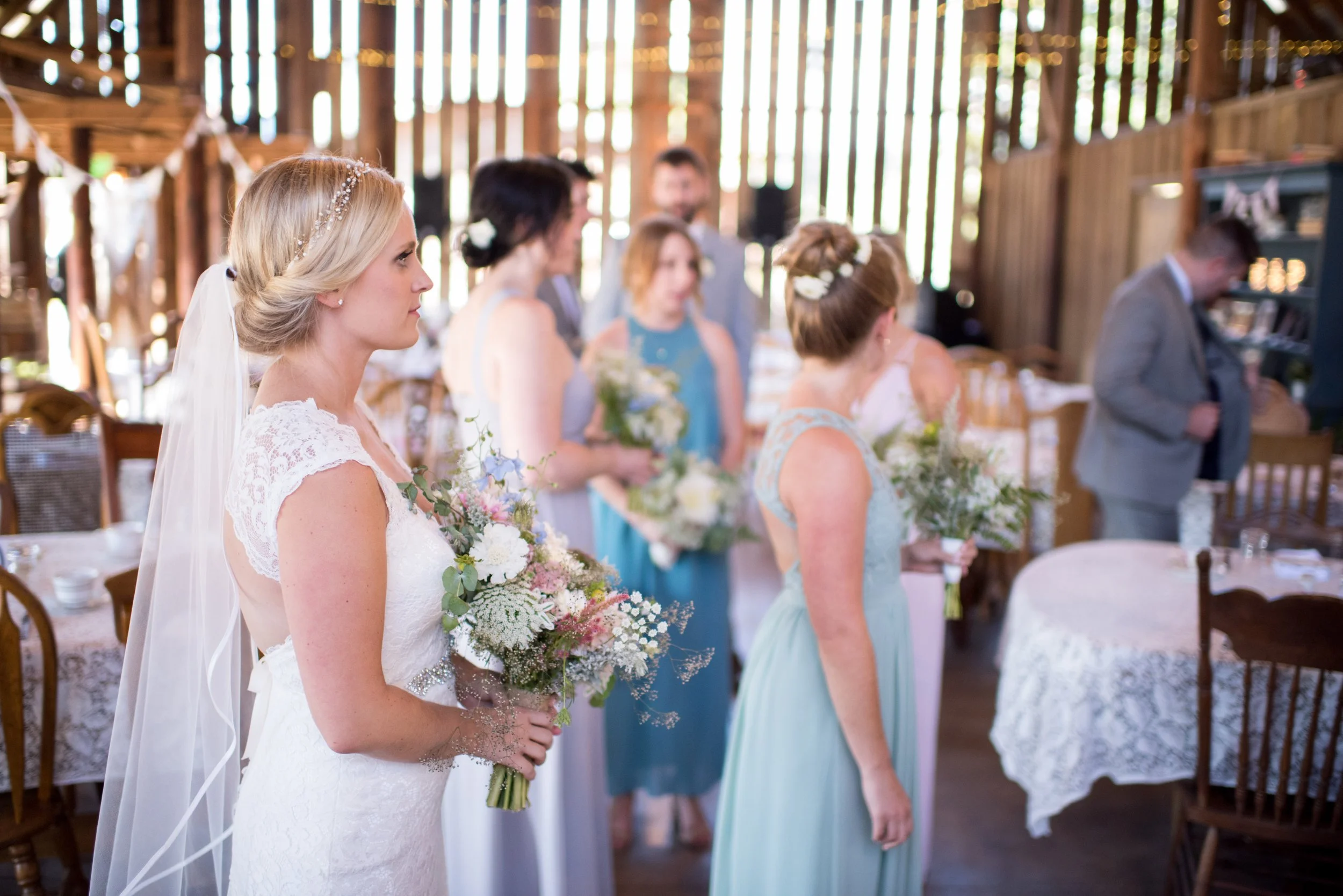 Bride in wedding dress holding bouquet during a ceremony in a rustic barn with bridesmaids and groomsmen.