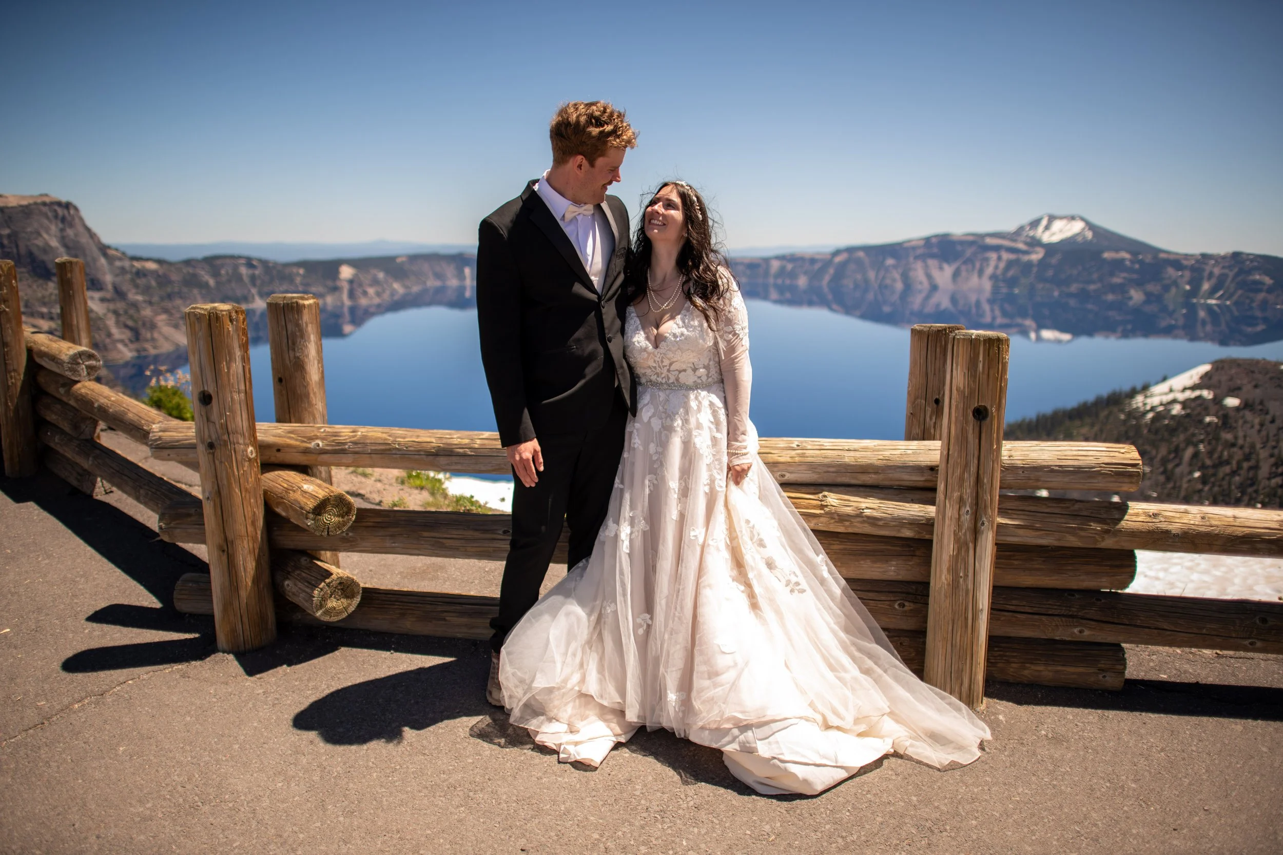 A newlywed couple dressed in wedding attire standing close and smiling at each other with a lake, mountains, and a clear blue sky in the background.