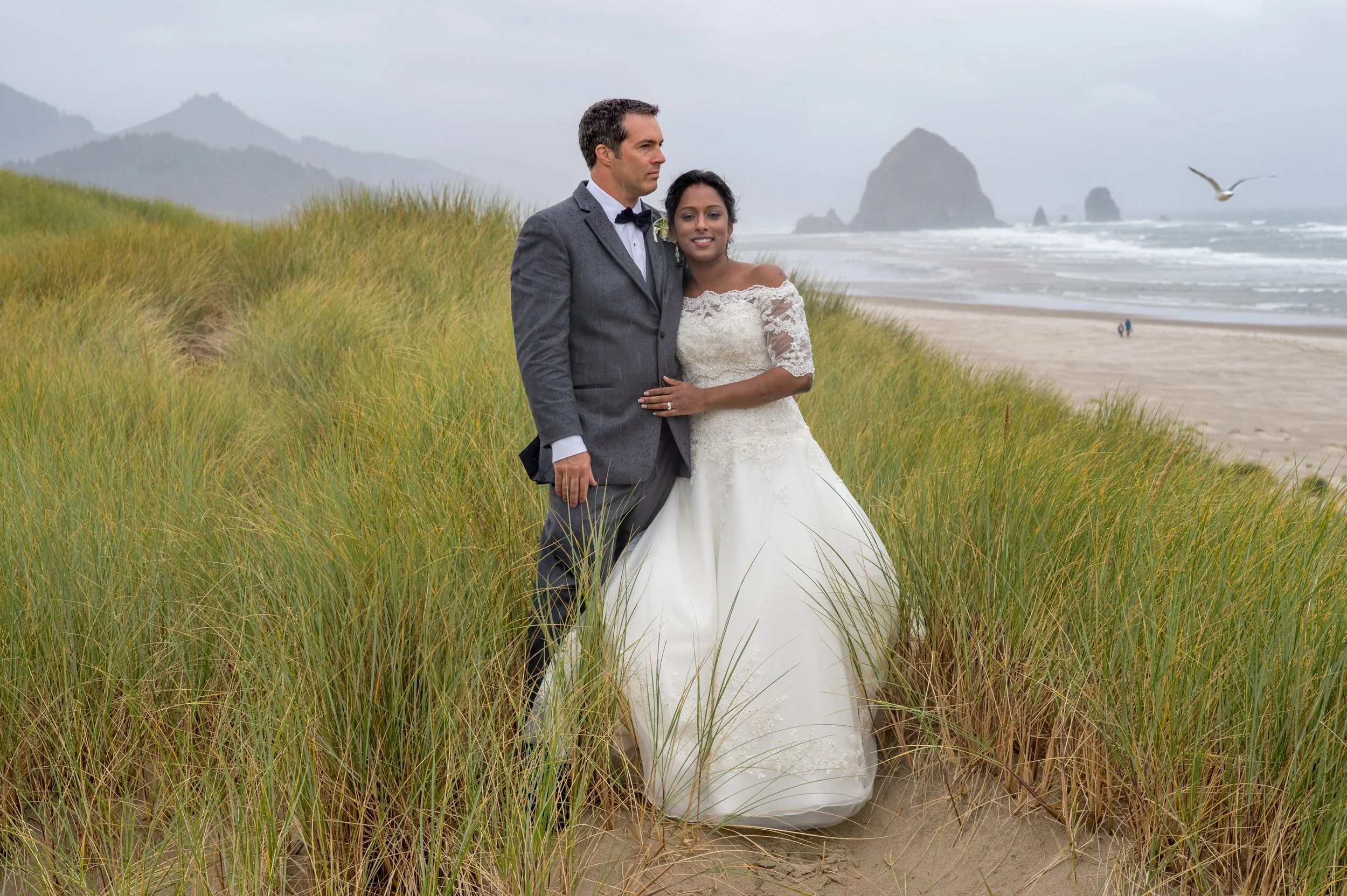 A bride and groom standing on a grassy sand dune by the beach on a cloudy day, with ocean waves, distant rock formations, and a seagull flying overhead.