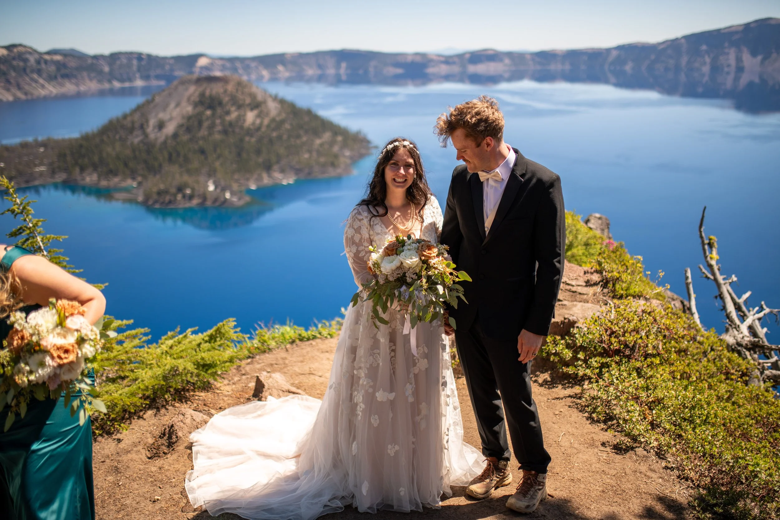 A bride and groom stand together during a wedding ceremony outdoors near a lake and mountains. The bride is holding a bouquet of flowers, wearing a white lace wedding dress, and the groom is in a black suit with a white shirt and bow tie. They are sm