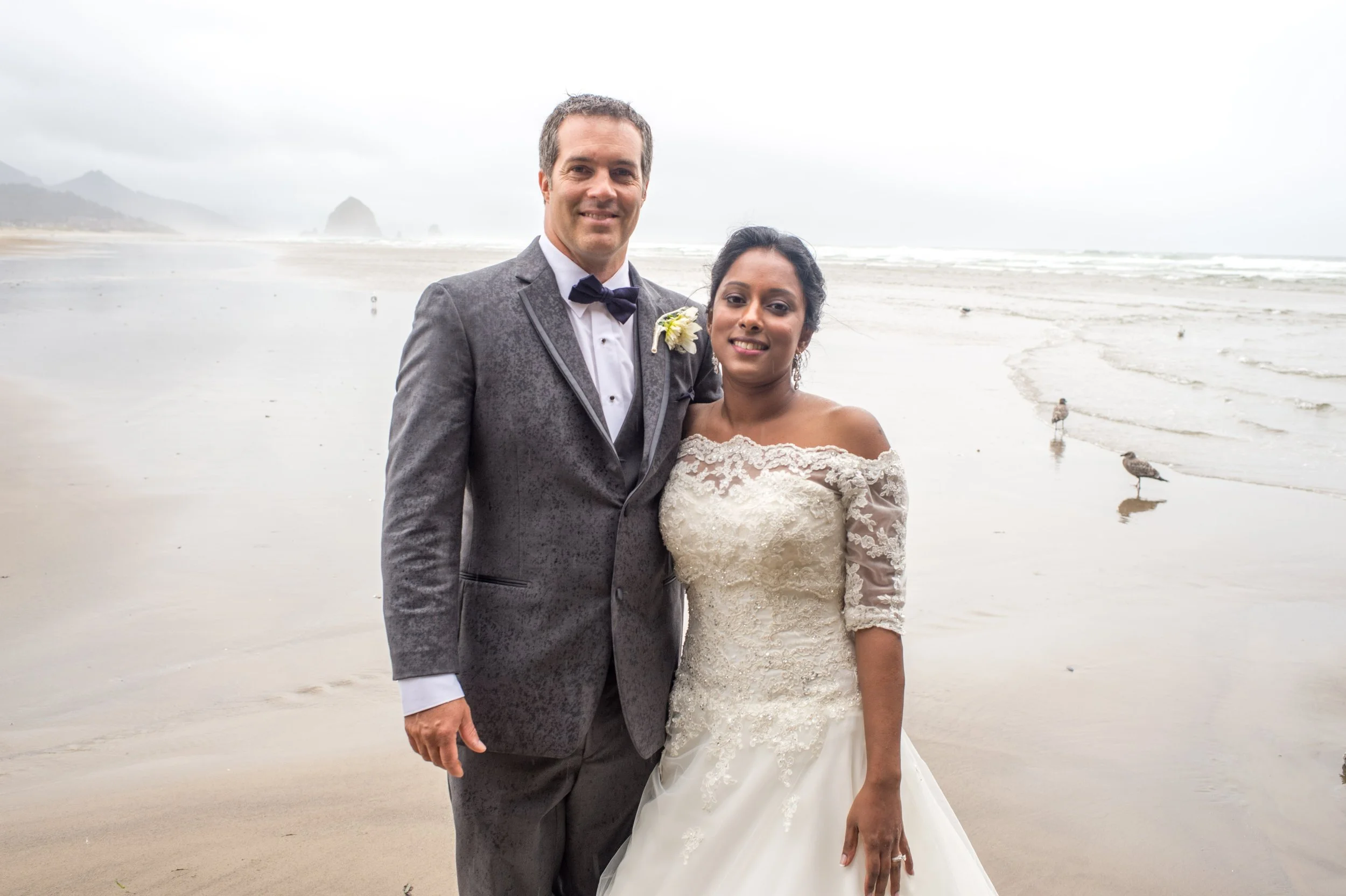 A newlywed couple, a man in a gray tuxedo with a black bow tie and a woman in a white wedding dress, standing on a beach with foggy weather, seagulls on the sand, and mountains in the background.