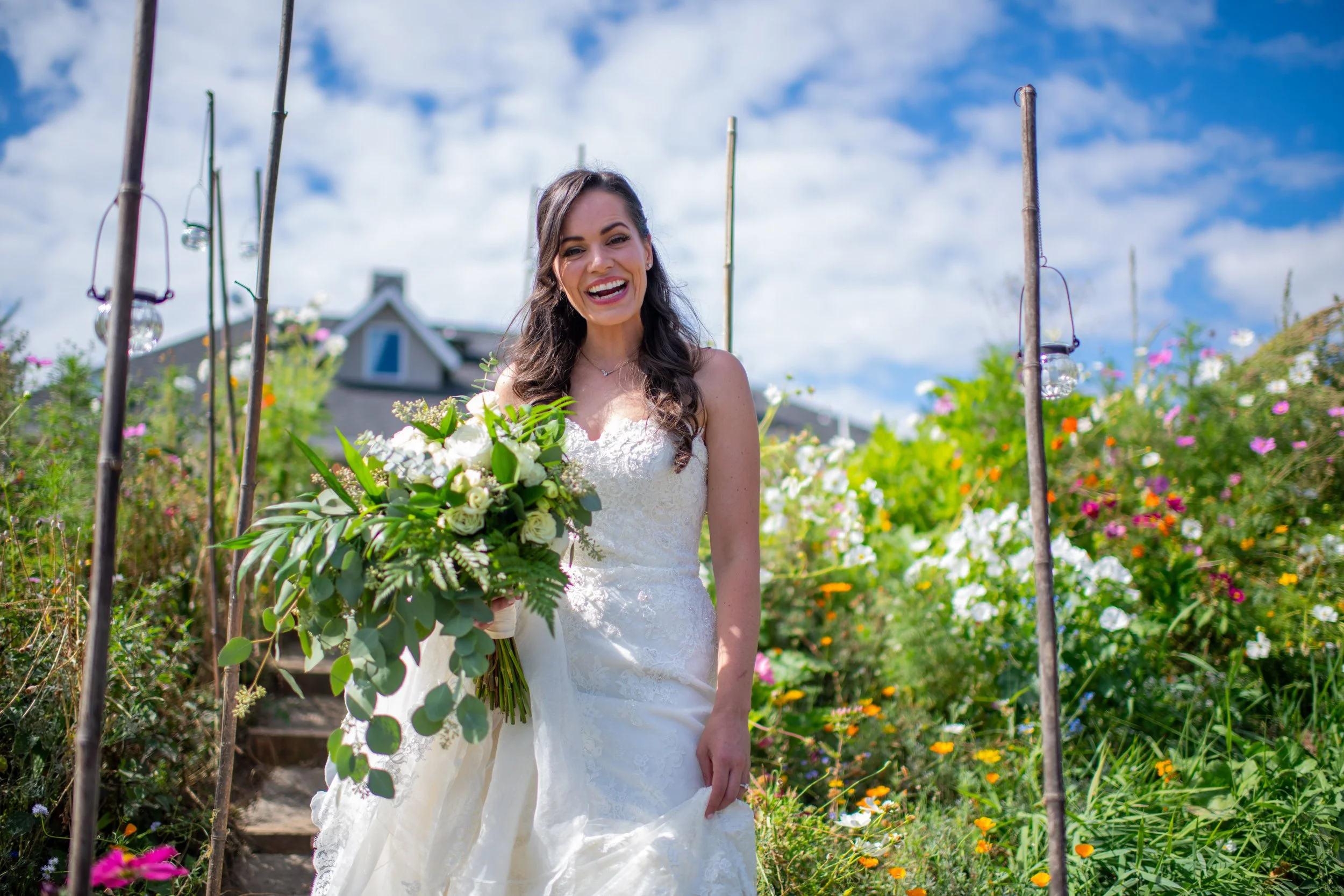 A smiling bride in a white wedding dress holding a bouquet of white and green flowers standing in a garden with colorful flowers and a house in the background.
