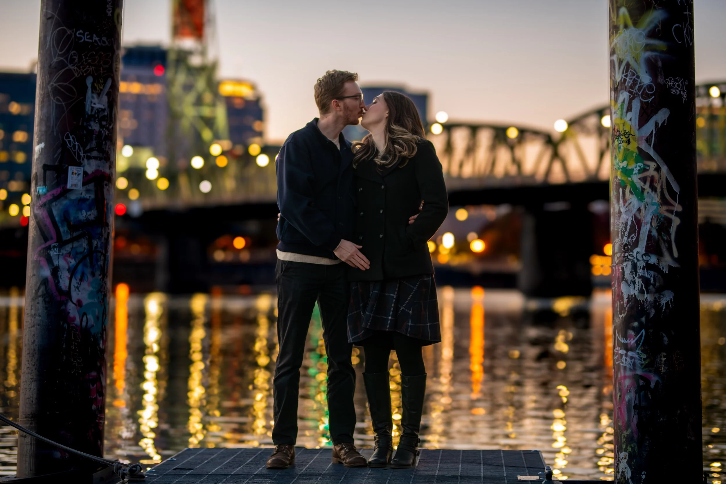 A couple kissing on a dock near the water at sunset, with blurred city skyline and bridge in the background, framed by two graffiti-covered poles.