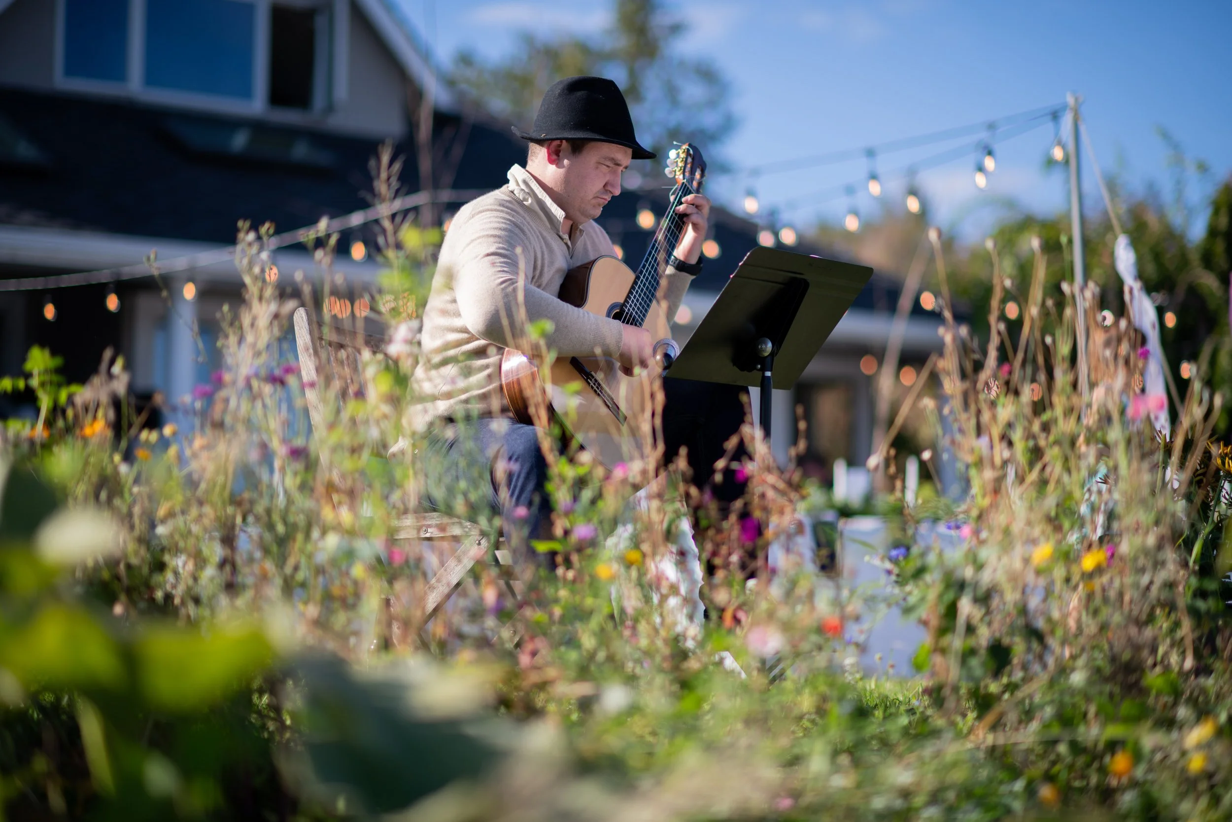 A man wearing a black hat and beige jacket playing an acoustic guitar outdoors while reading sheet music, with string lights hanging in the background and flowers in the foreground.