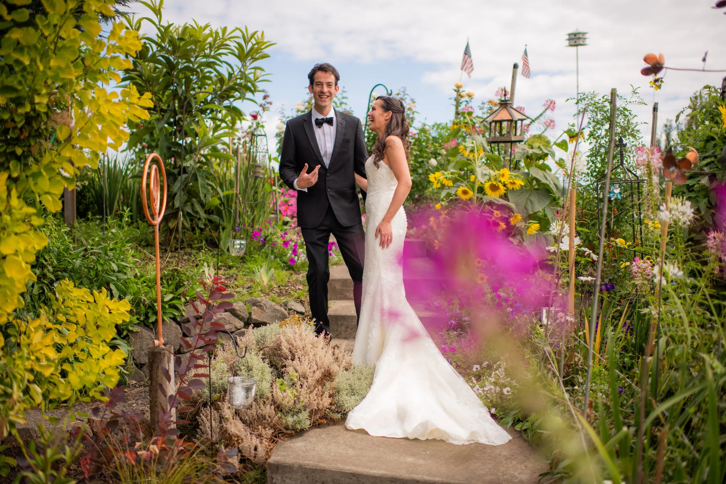 A happy couple, dressed in wedding attire, walking in a colorful garden with flowers and plants, during a bright day with partly cloudy skies.