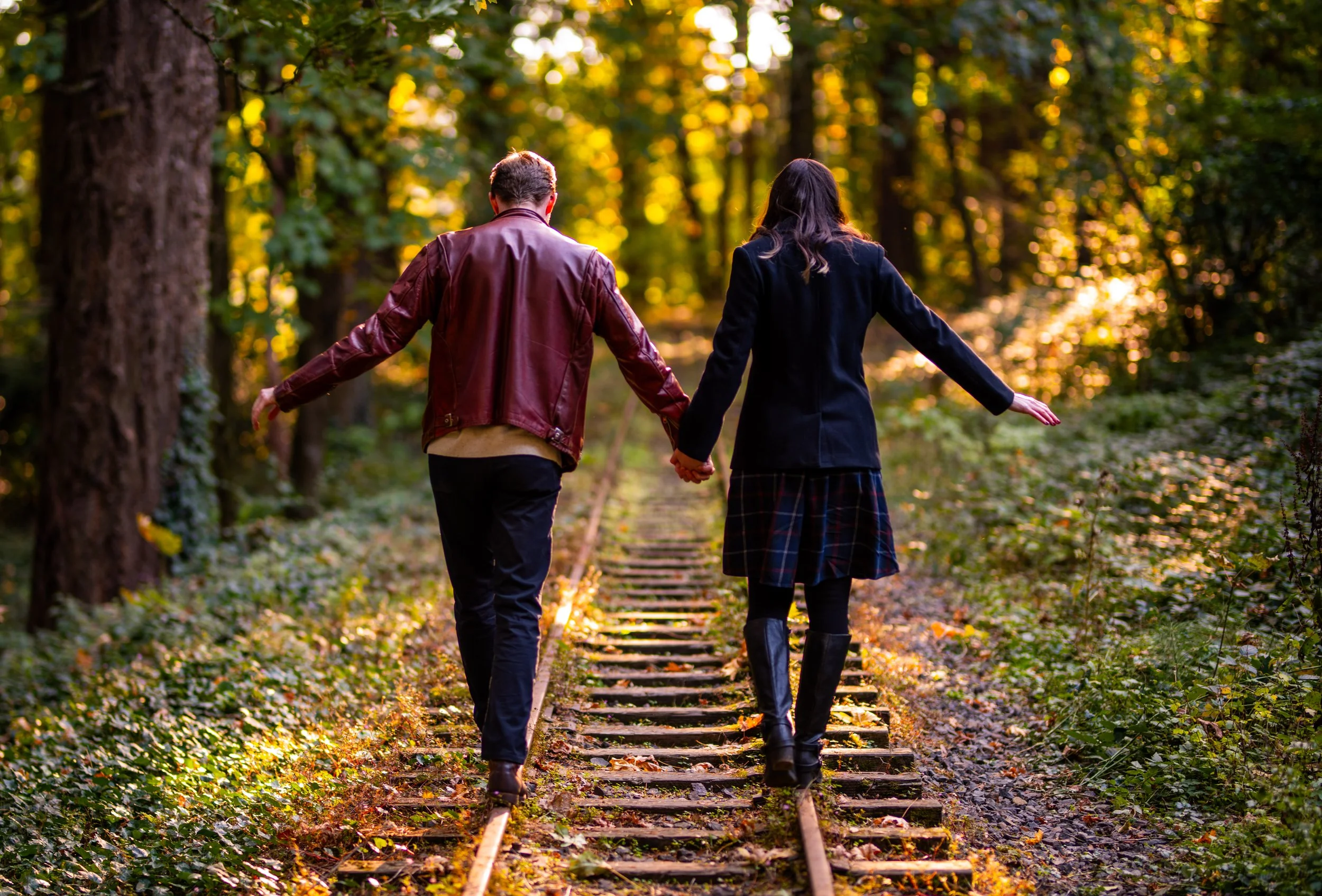 A couple holding hands and walking down a set of railroad tracks through a wooded area during fall, with sunlight filtering through the trees.