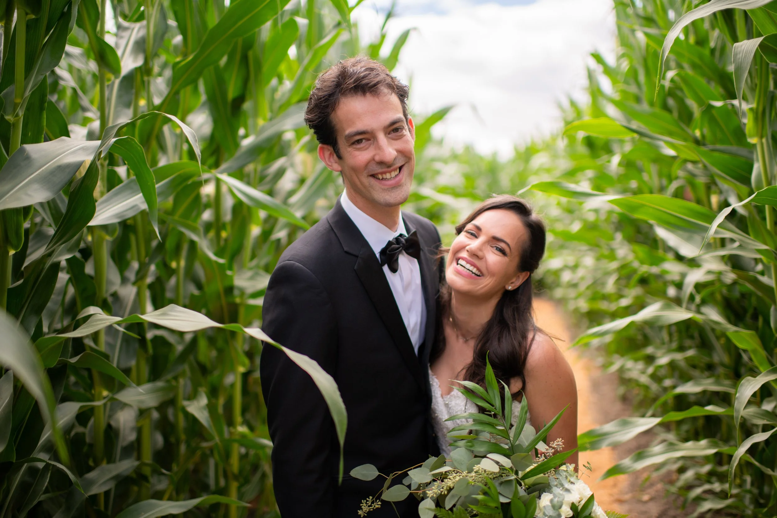 A smiling couple in wedding attire, a man in a tuxedo and woman in a wedding dress, standing in a lush green cornfield.