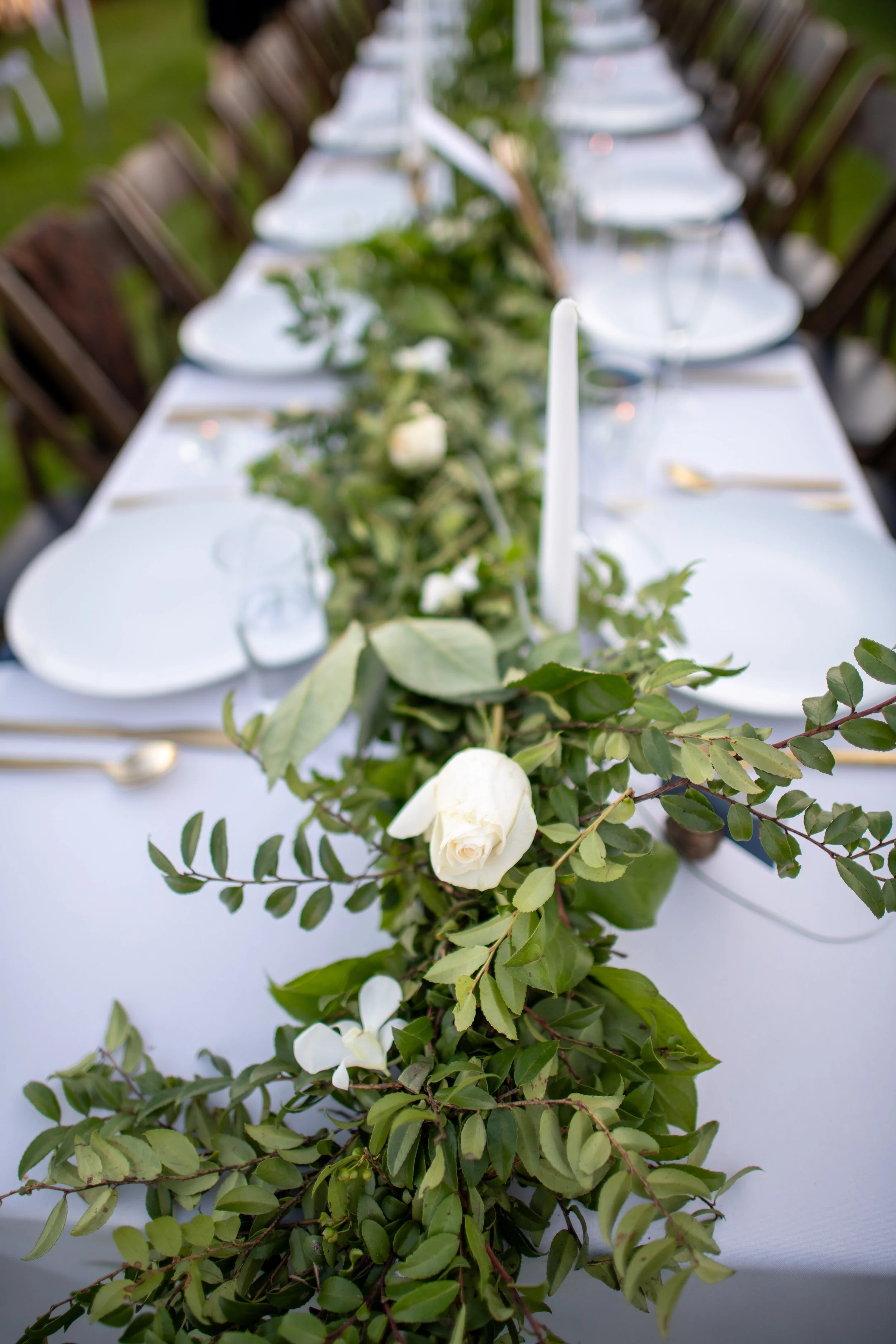 A close-up of a table decorated with green leaves and white flowers, with white plates, glasses, and gold utensils, set outdoors for a formal event.