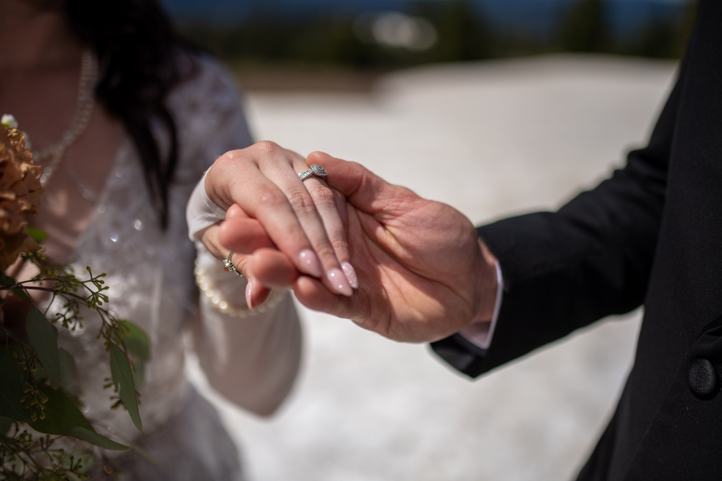 Close-up of a bride and groom holding hands during a wedding ceremony, focusing on the bride's diamond ring and wedding gown on a sunny outdoor setting.