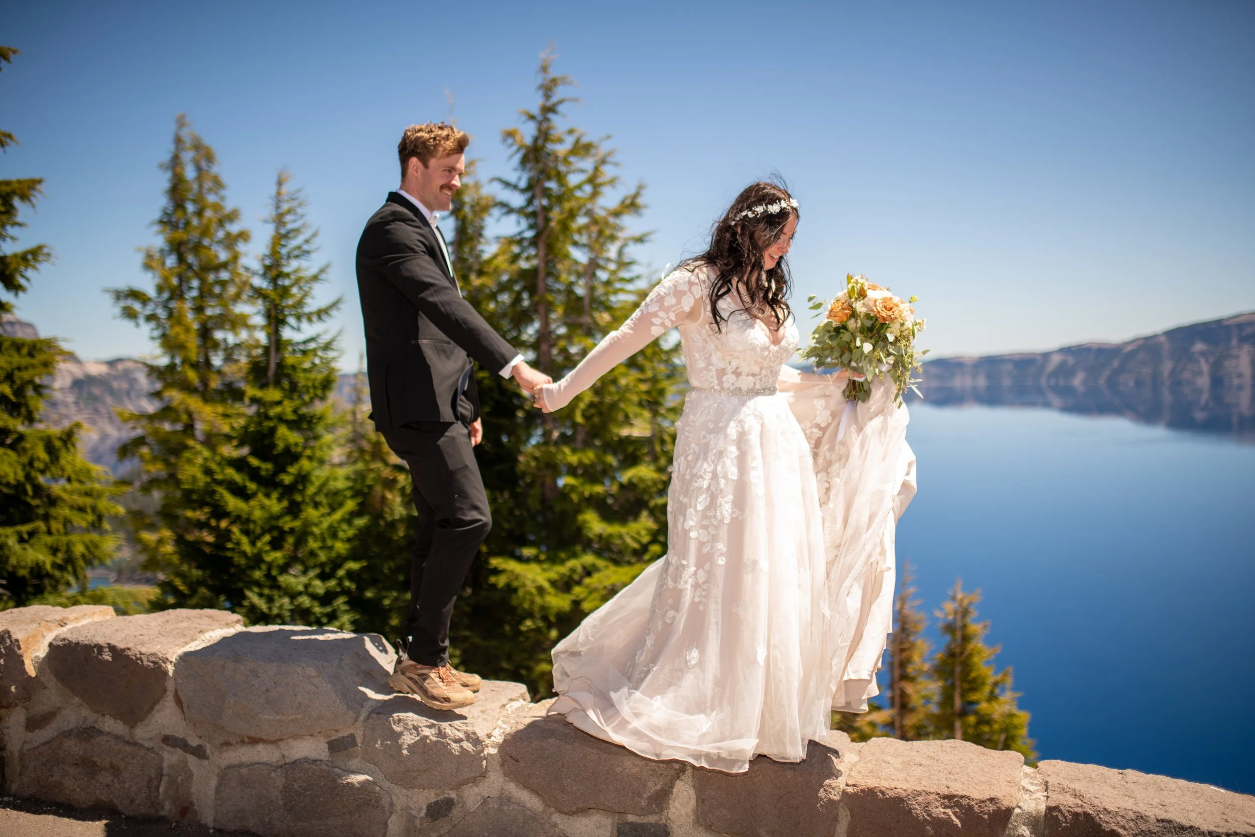 A bride and groom holding hands and walking together outdoors near a lake, with green trees and mountains in the background on a clear, sunny day.
