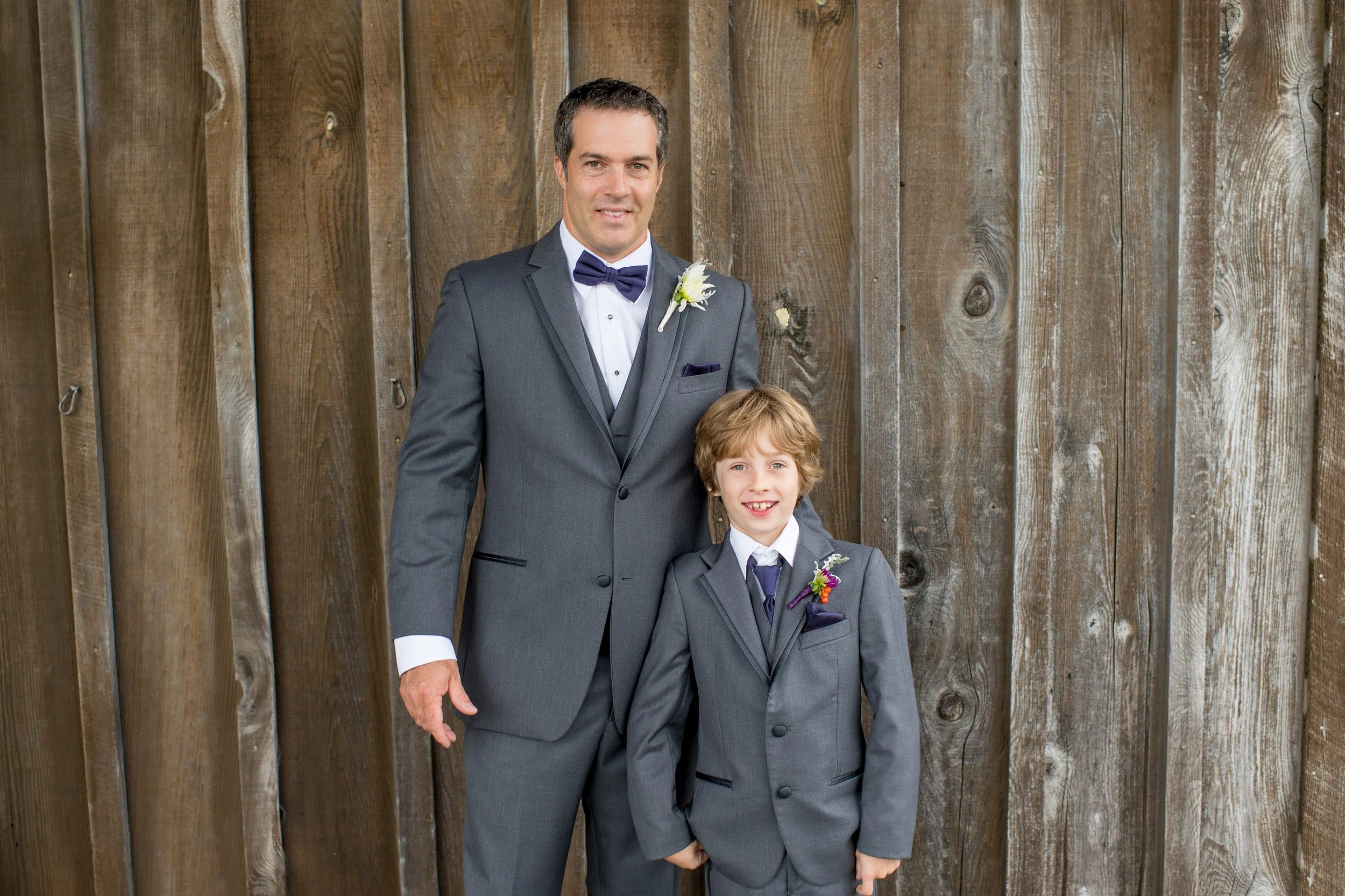 A man in a gray suit with a bow tie and white dress shirt standing beside a young boy in a matching gray suit with a purple tie, both smiling in front of a wooden fence.