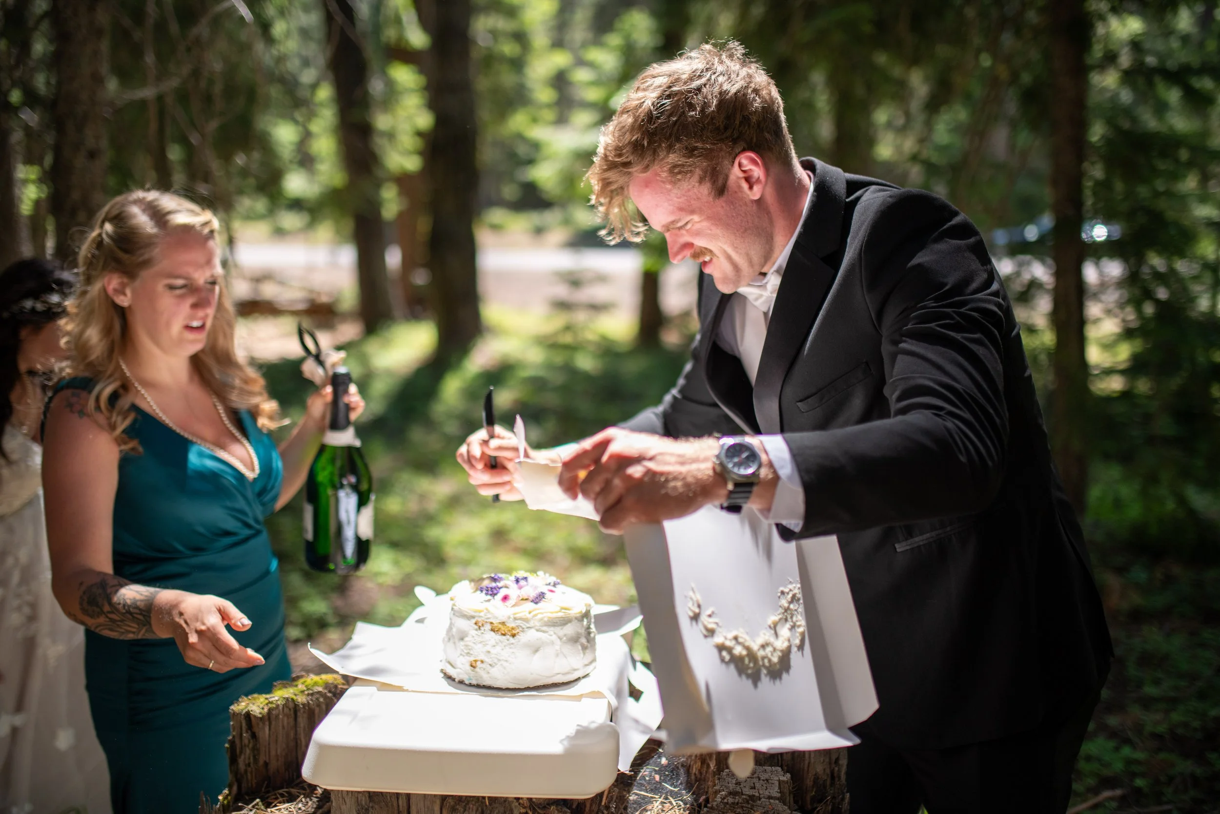 A man in a black blazer signing a paper at an outdoor celebration table with a white cake, a woman in a blue dress holding a bottle of champagne, and trees around.