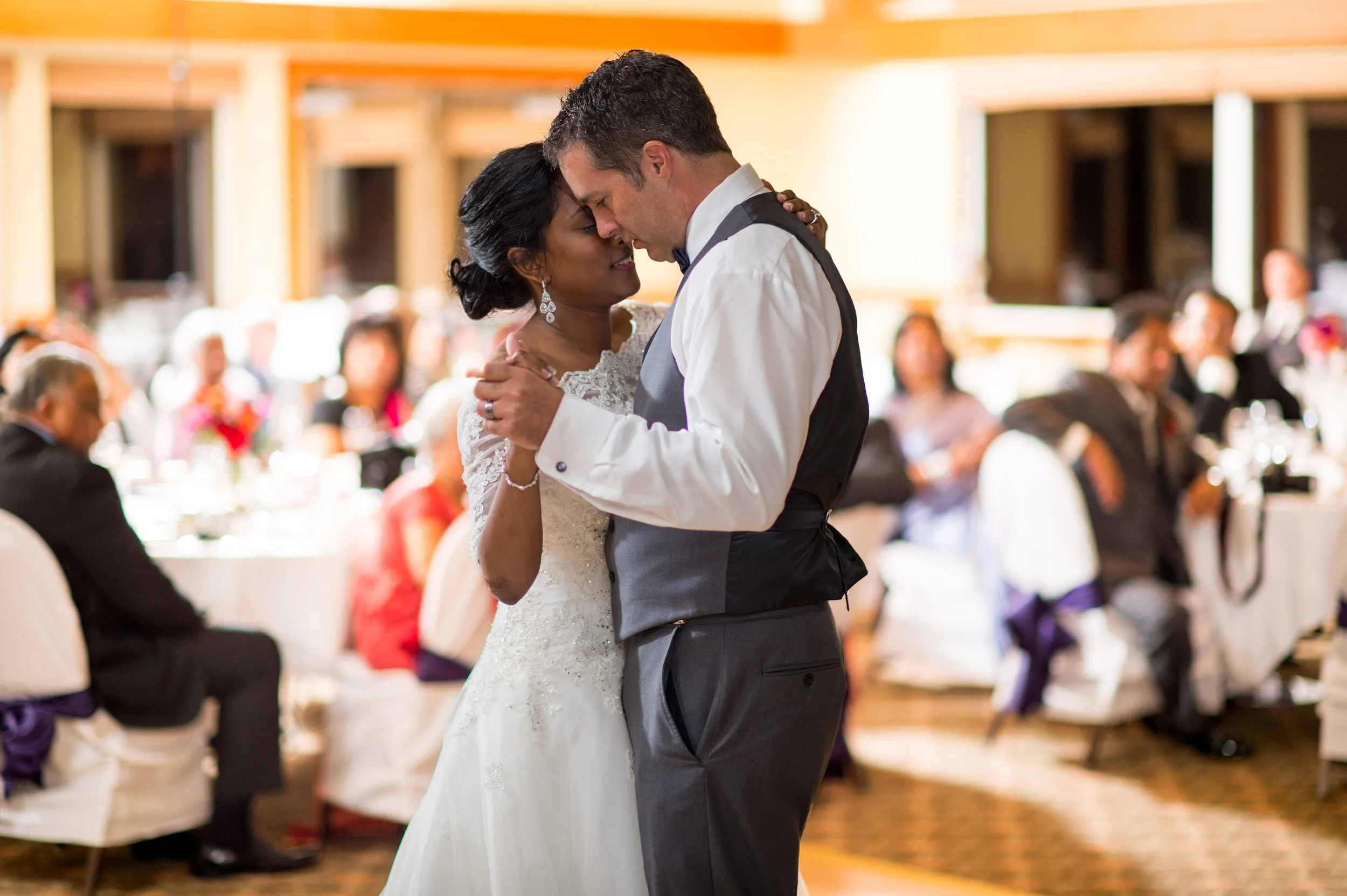A bride and groom share a dance at their wedding reception, with guests seated at tables in the background.