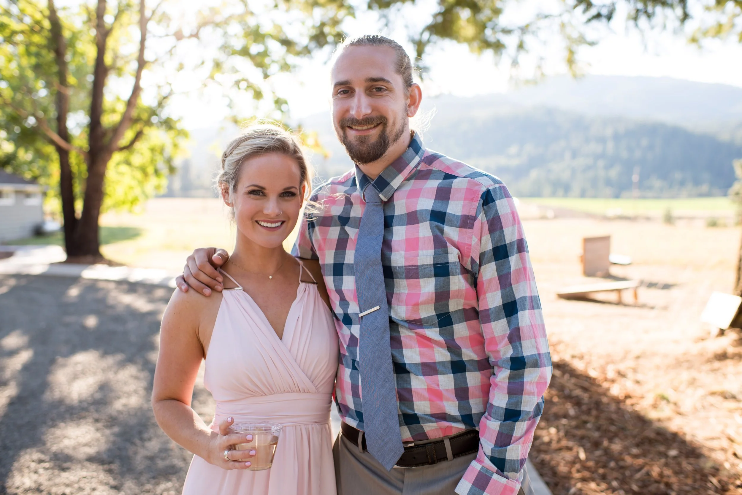 A smiling man and woman standing outdoors in a park with trees and mountains in the background. The woman is holding a glass of wine. The man has long hair tied back and is wearing a plaid shirt and blue tie. The woman has blonde hair and is wearing 
