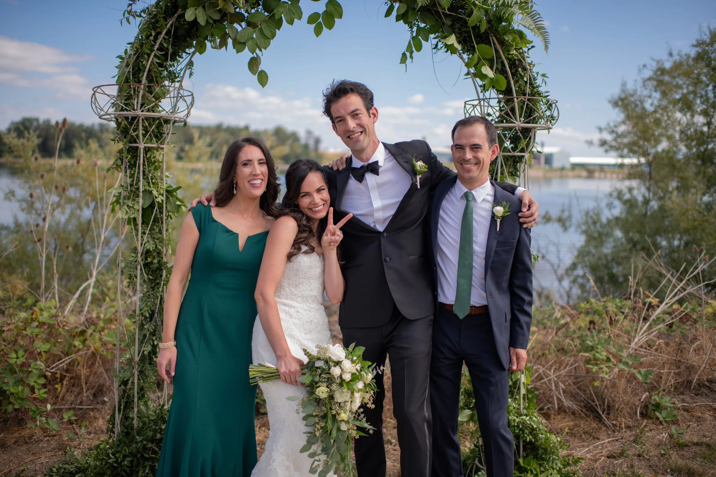 A group of five people, including a bride in a white dress holding a bouquet of white roses and four guests, standing under a decorative archway outdoors. The landscape features a lake, trees, and a blue sky with clouds.