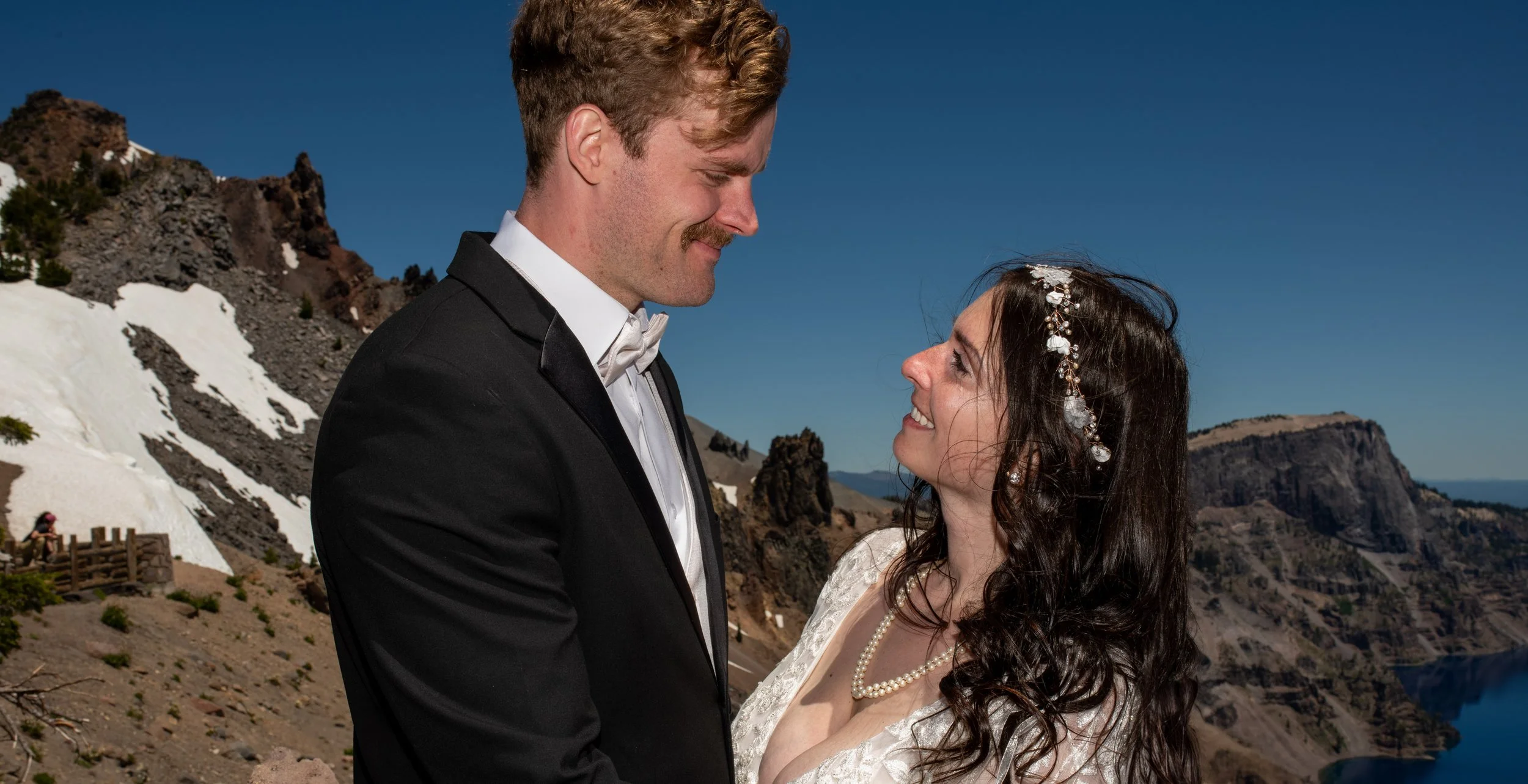 A couple dressed in wedding attire outdoors, smiling and looking at each other against a mountainous backdrop with snow patches and rocky formations.