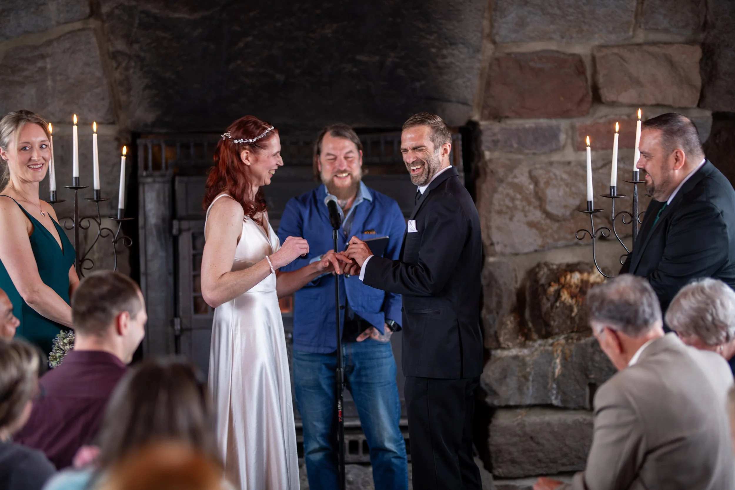 A wedding ceremony taking place indoors with five people standing at the altar, exchanging vows, amid an audience of seated guests. The groom and bride are smiling, with the officiant standing between them. The setting features stone walls and candel