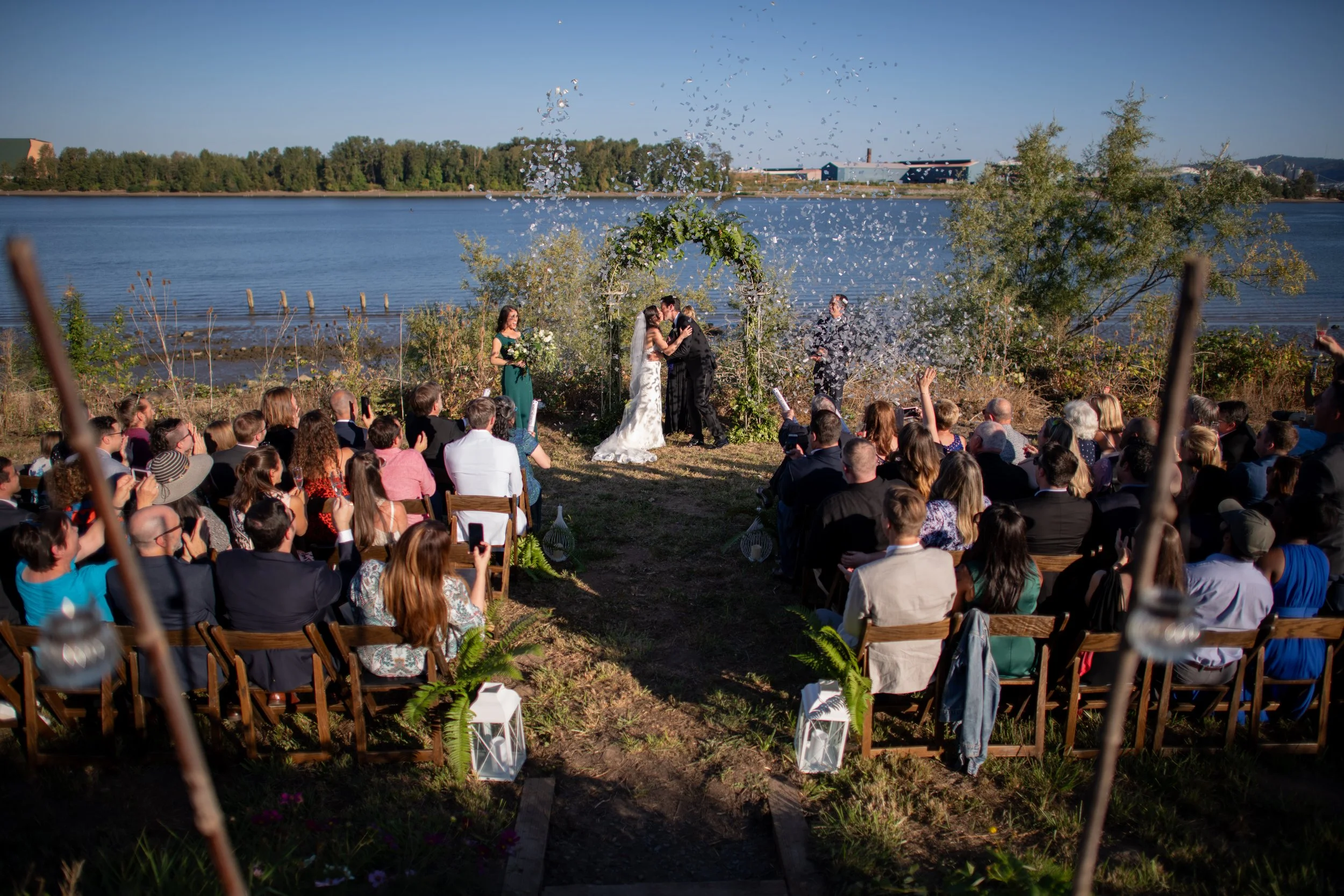 A large outdoor wedding ceremony near a body of water with a bride and groom kissing under an arch, surrounded by guests seated in rows, and a officiant standing nearby. Confetti is falling from the sky, and trees and industrial buildings are visible
