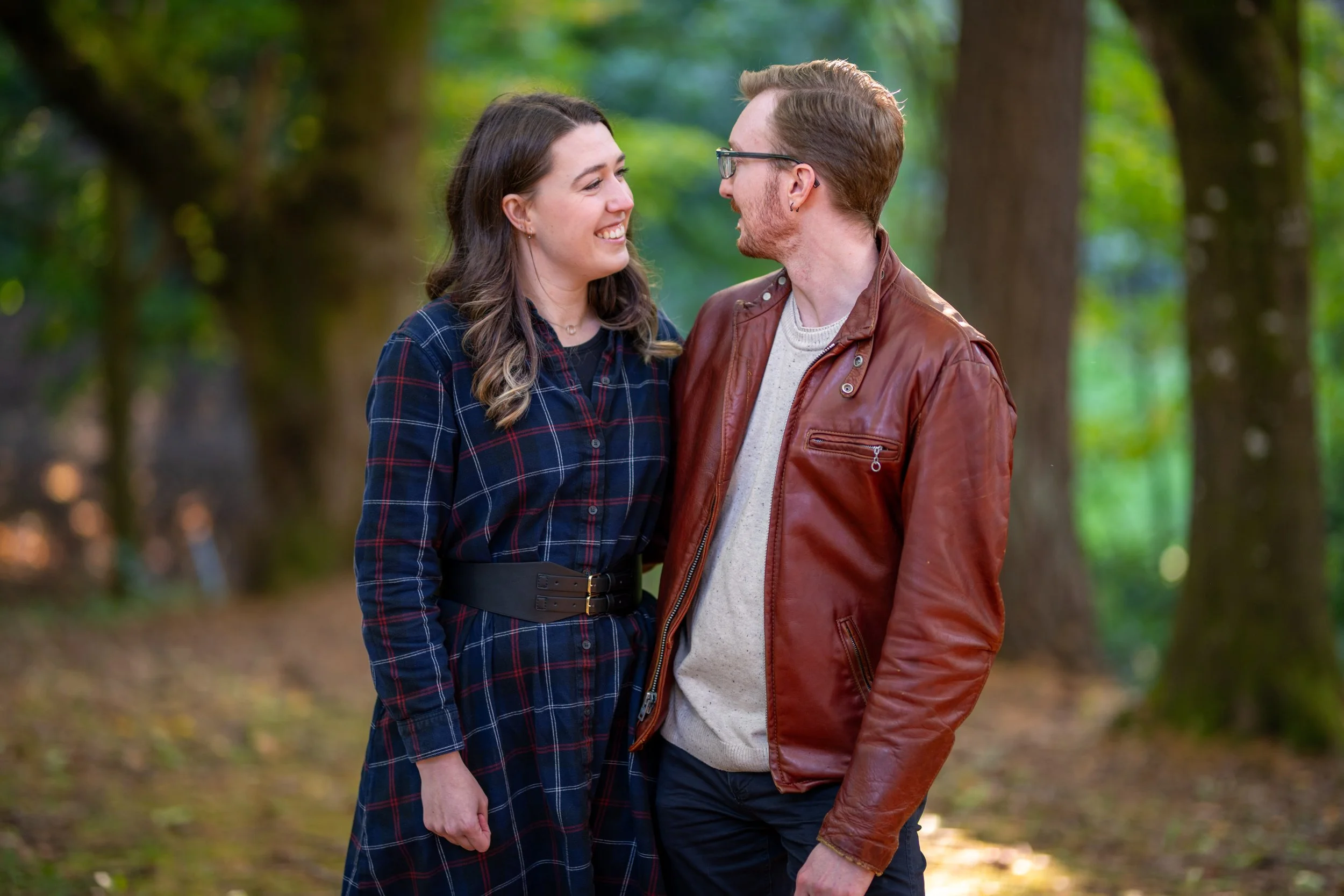 A young couple standing close together outdoors, smiling and looking into each other's eyes, in a park with trees in the background.