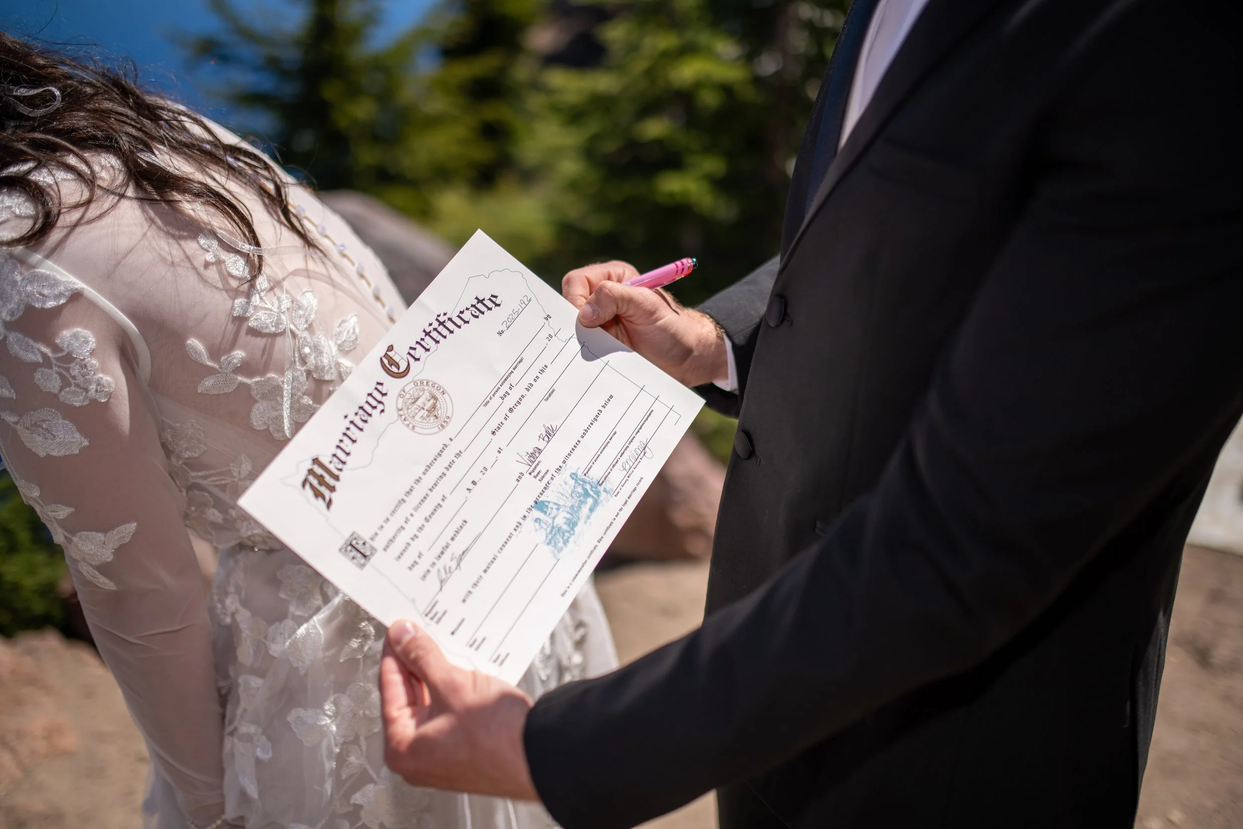 A person in a dark suit signing a marriage certificate with a pink pen during an outdoor wedding ceremony.