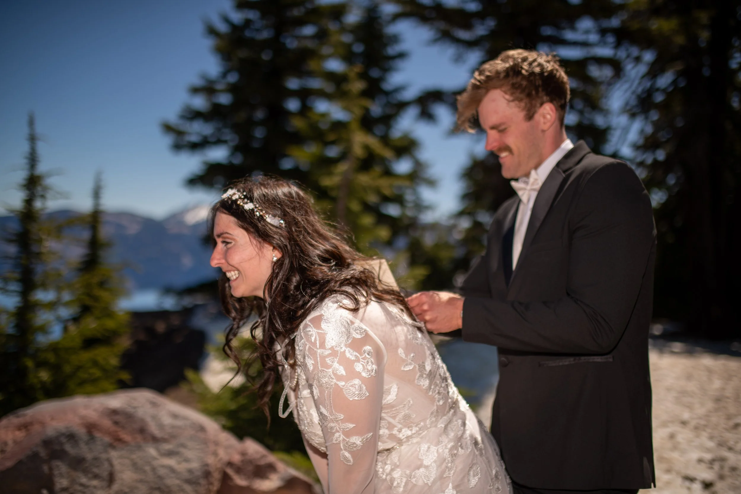A bride and groom are standing outdoors in a forested area with mountains in the background. The bride has long, curly hair and is wearing a lace wedding dress with a floral headpiece. The groom is wearing a black tuxedo and white bow tie. They are s