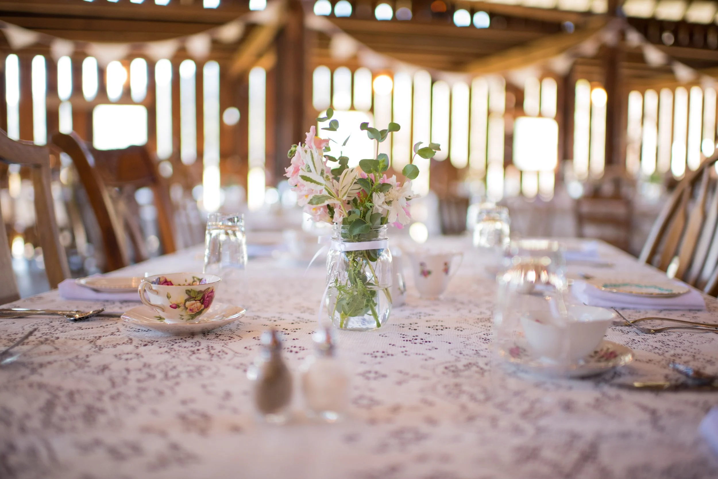 A table set for a celebration or gathering with a floral centerpiece, teacups, and glassware on a lace tablecloth in a rustic wooden venue with string lights.