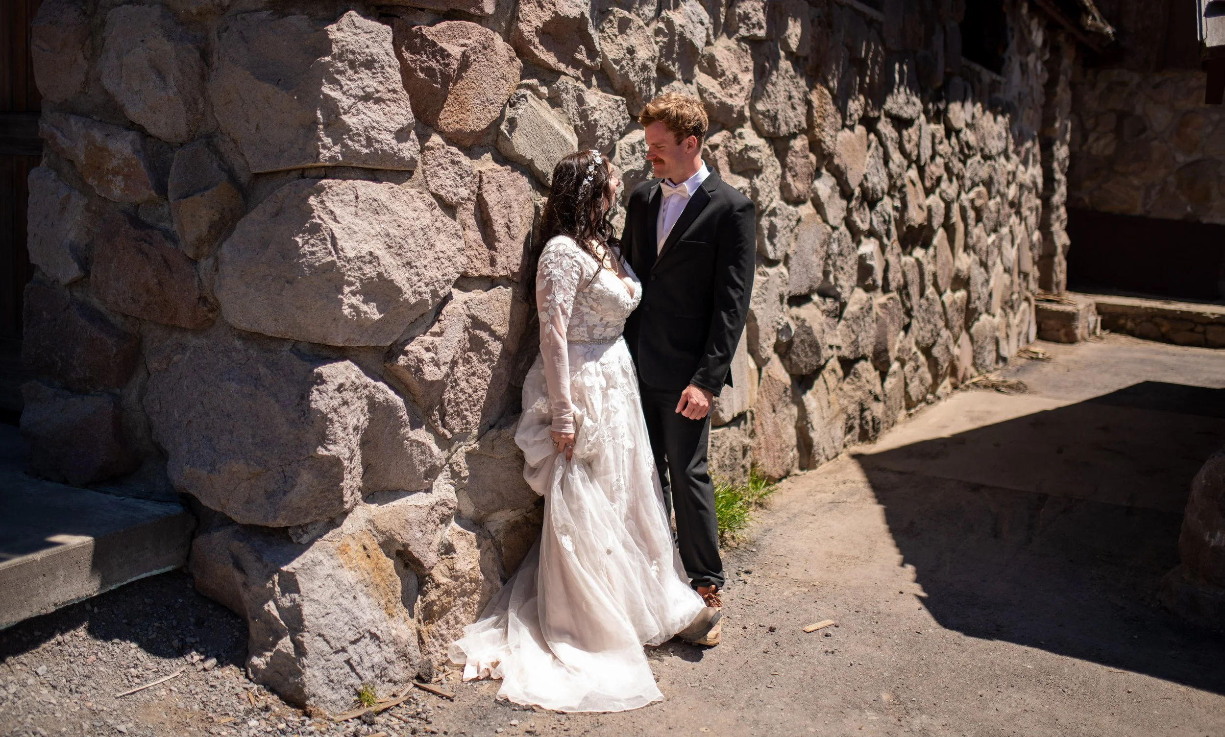 A bride and groom standing close together outdoors on a sunny day, with the bride leaning against a large stone wall, both looking into each other's eyes, dressed in wedding attire.