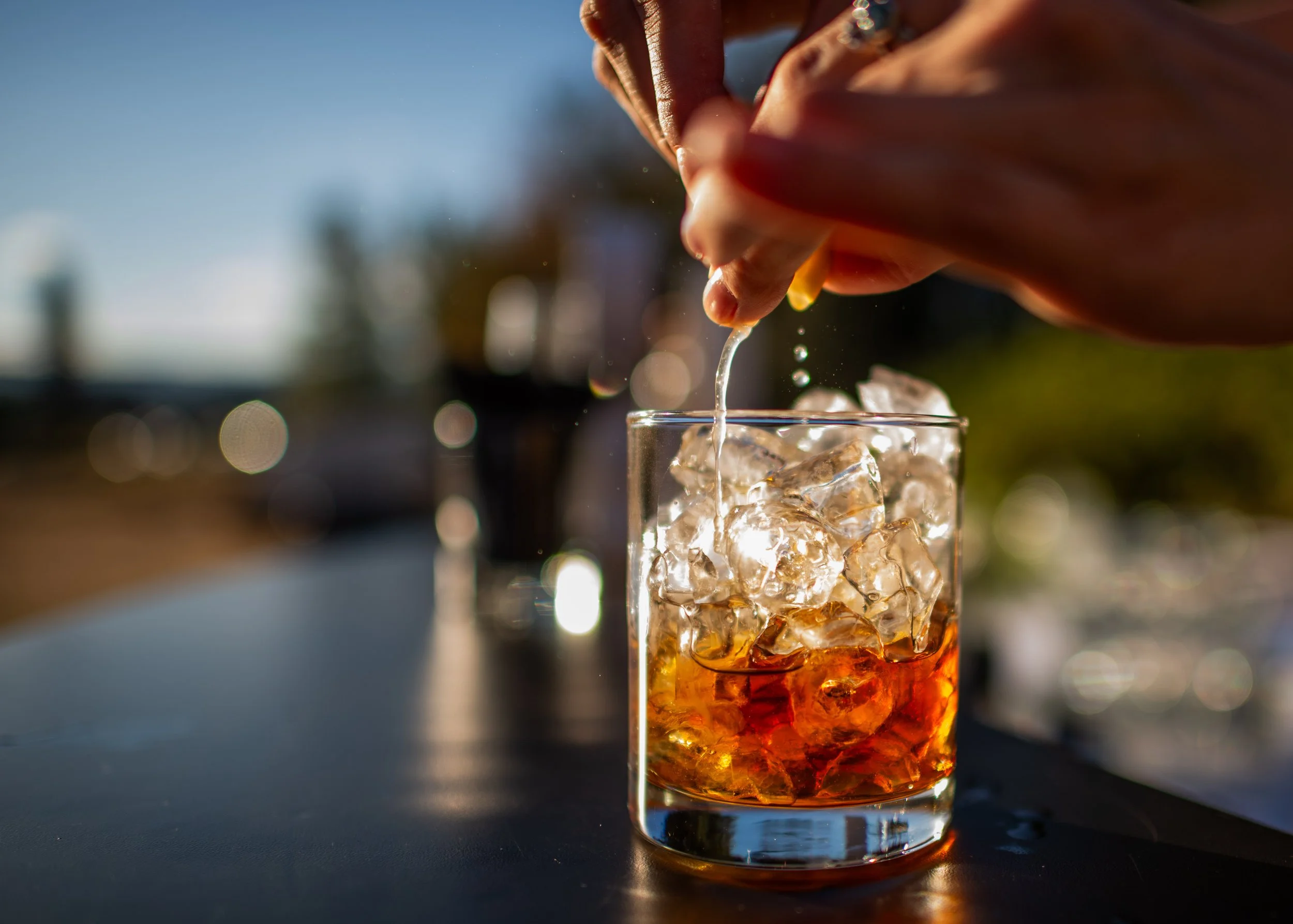 Close-up of a hand dropping ice cubes into a glass filled with a dark-colored beverage outdoors on a sunny day.