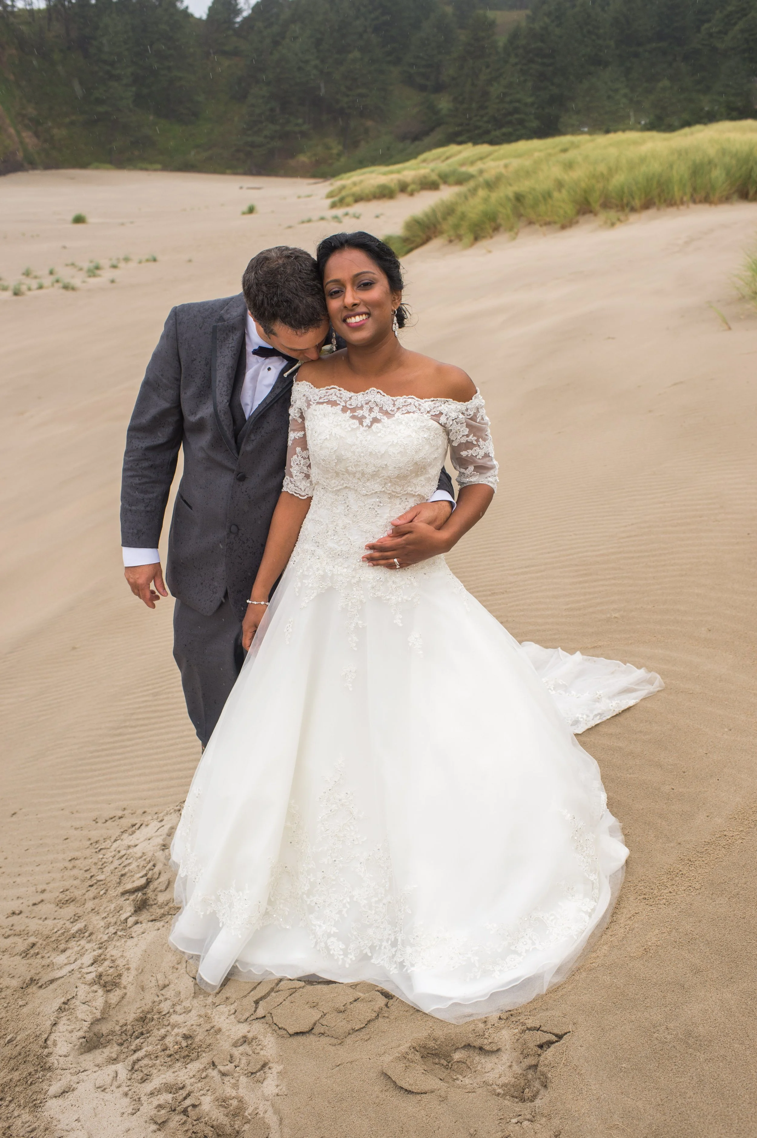 A bride in a white lace wedding dress and a groom in a tuxedo hugging and smiling on a sandy beach with dune grass and greenery in the background.
