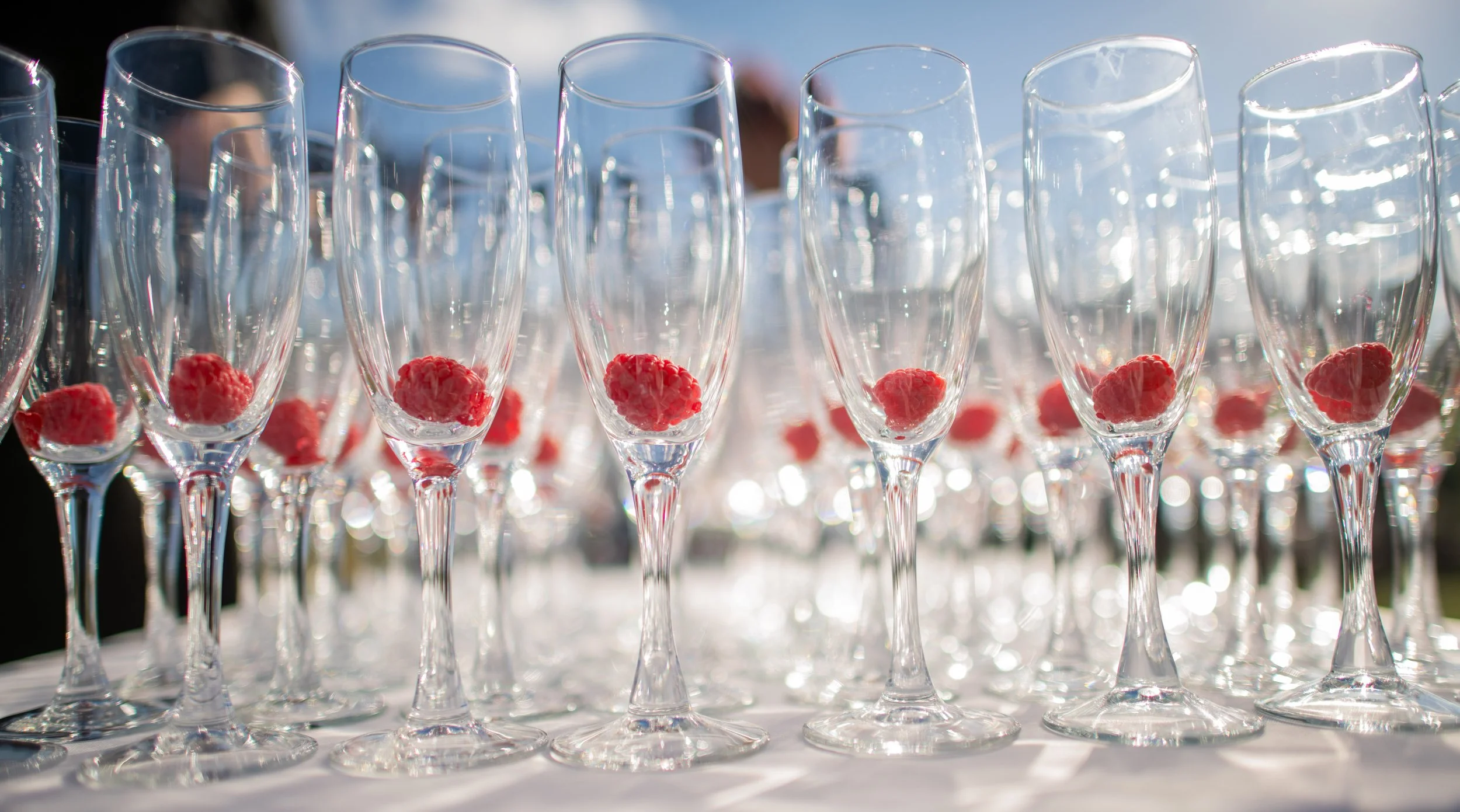 Multiple empty champagne flutes with pink flowers in the bottom, arranged outdoors on a white table with bright sunlight and sky in the background.
