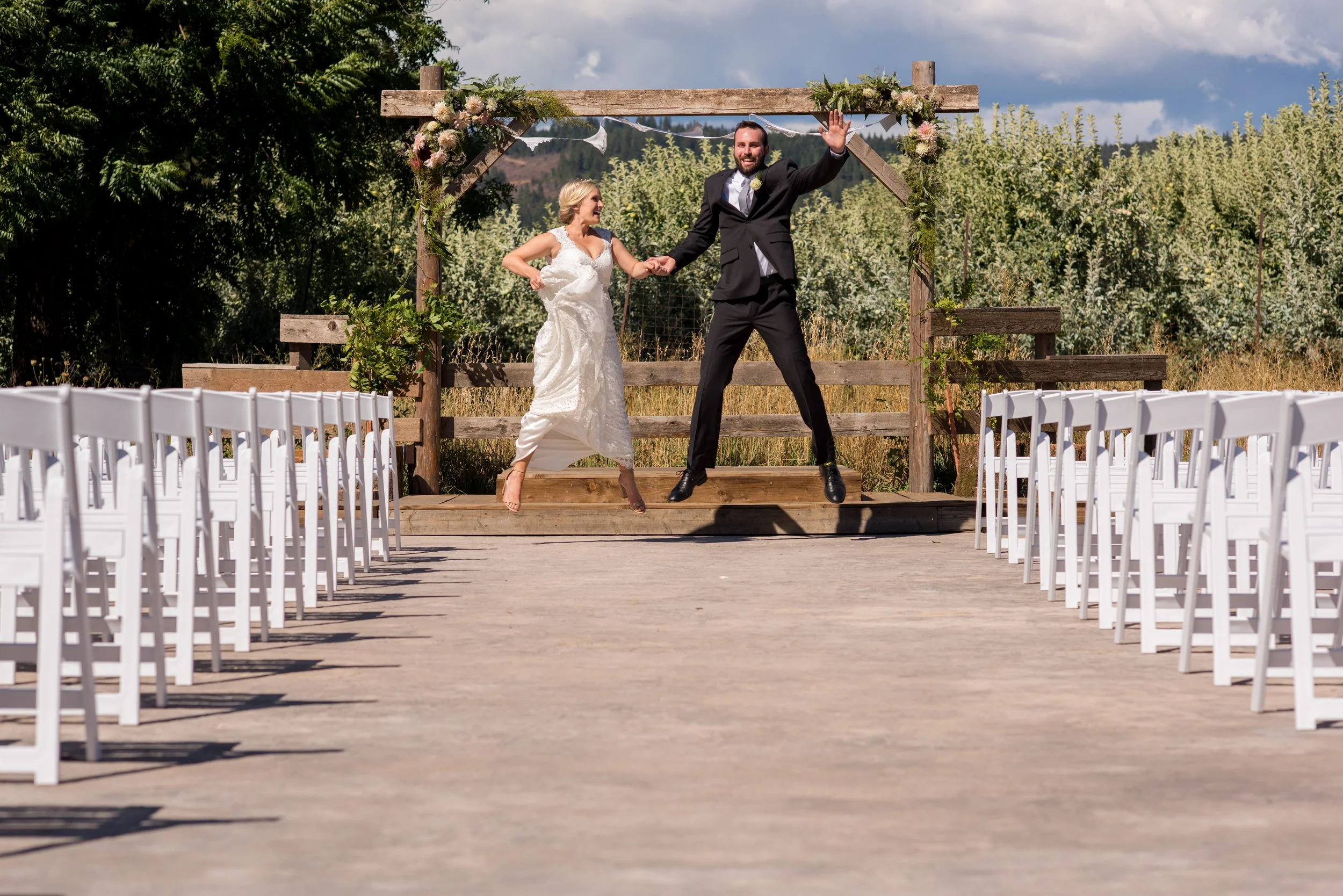 A couple dressed in wedding attire holding hands and smiling on a wooden platform at an outdoor wedding ceremony. There are rows of white chairs on either side and a rustic wooden arch decorated with flowers and greenery behind them. The background f