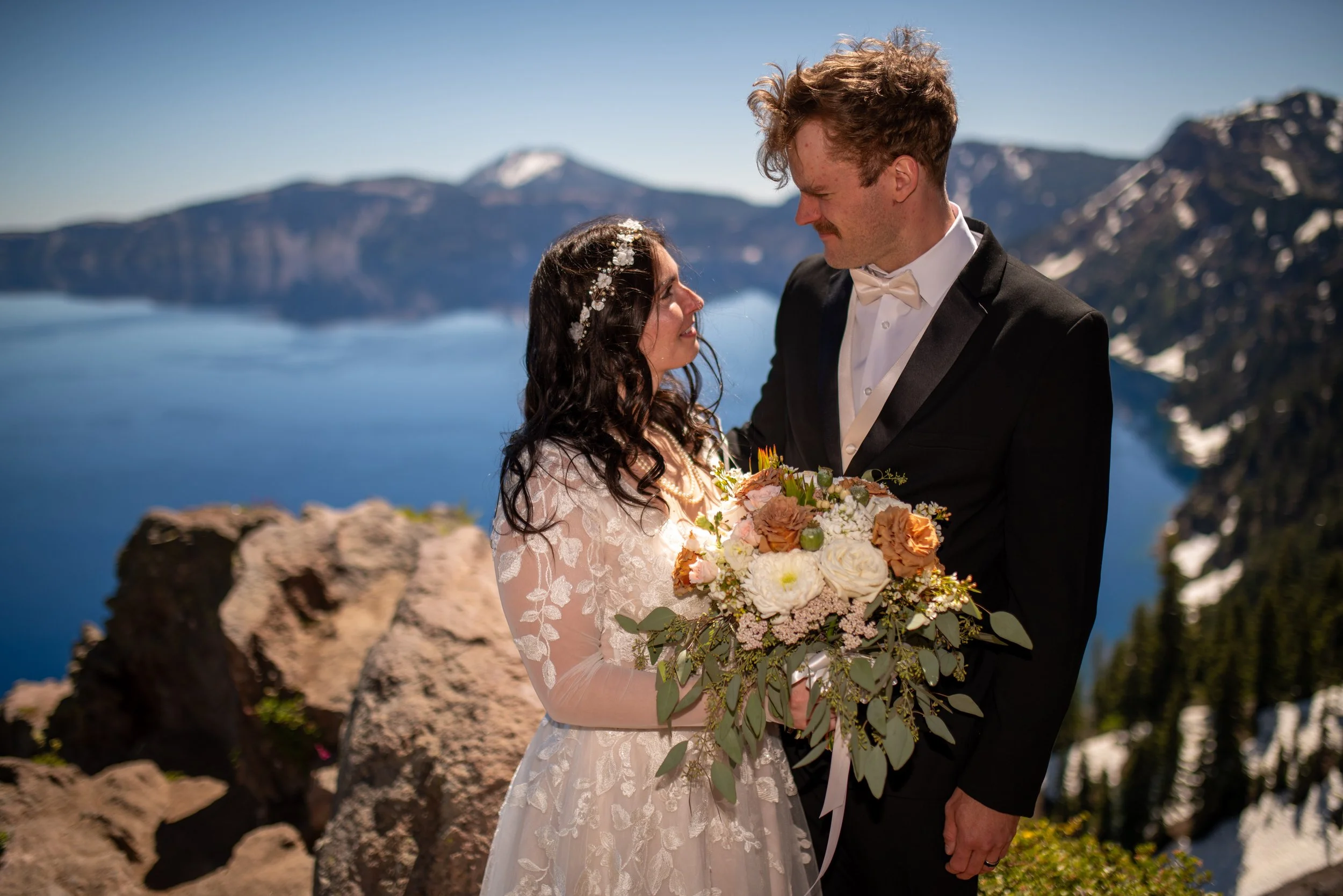 A bride and groom on their wedding day, standing outdoors near a body of water with mountains in the background. The bride is holding a large bouquet of flowers, and the groom is dressed in a tuxedo with a bow tie, looking lovingly at each other.