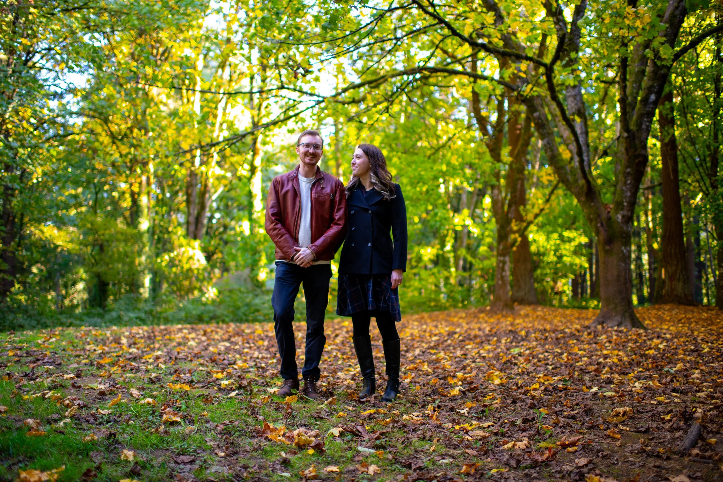 A smiling man and woman walking in a park with fall foliage, yellow leaves on the trees and ground.