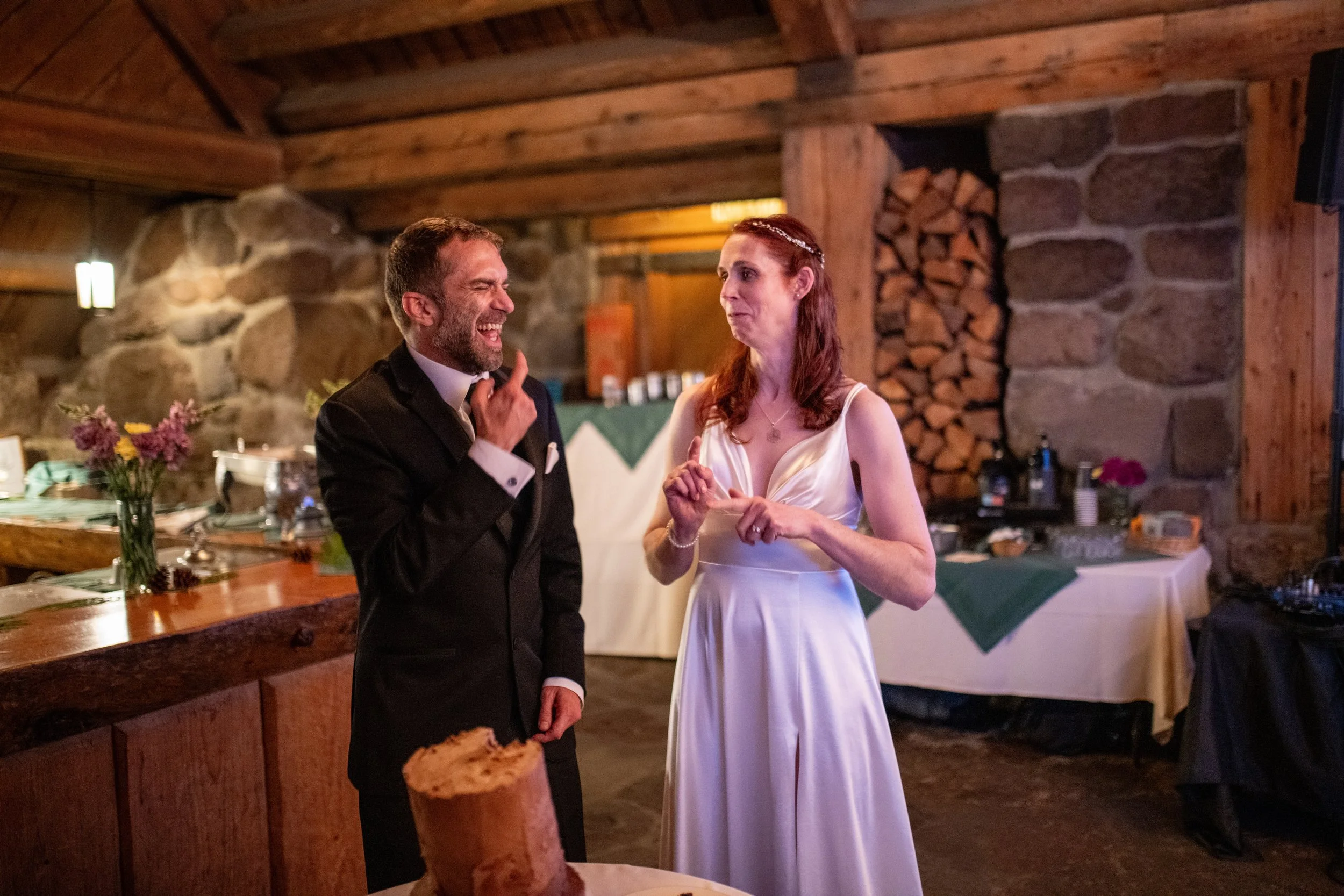 A groom in a black tuxedo and a bride in a white wedding gown are dancing and smiling at each other in a rustic wooden wedding reception hall.