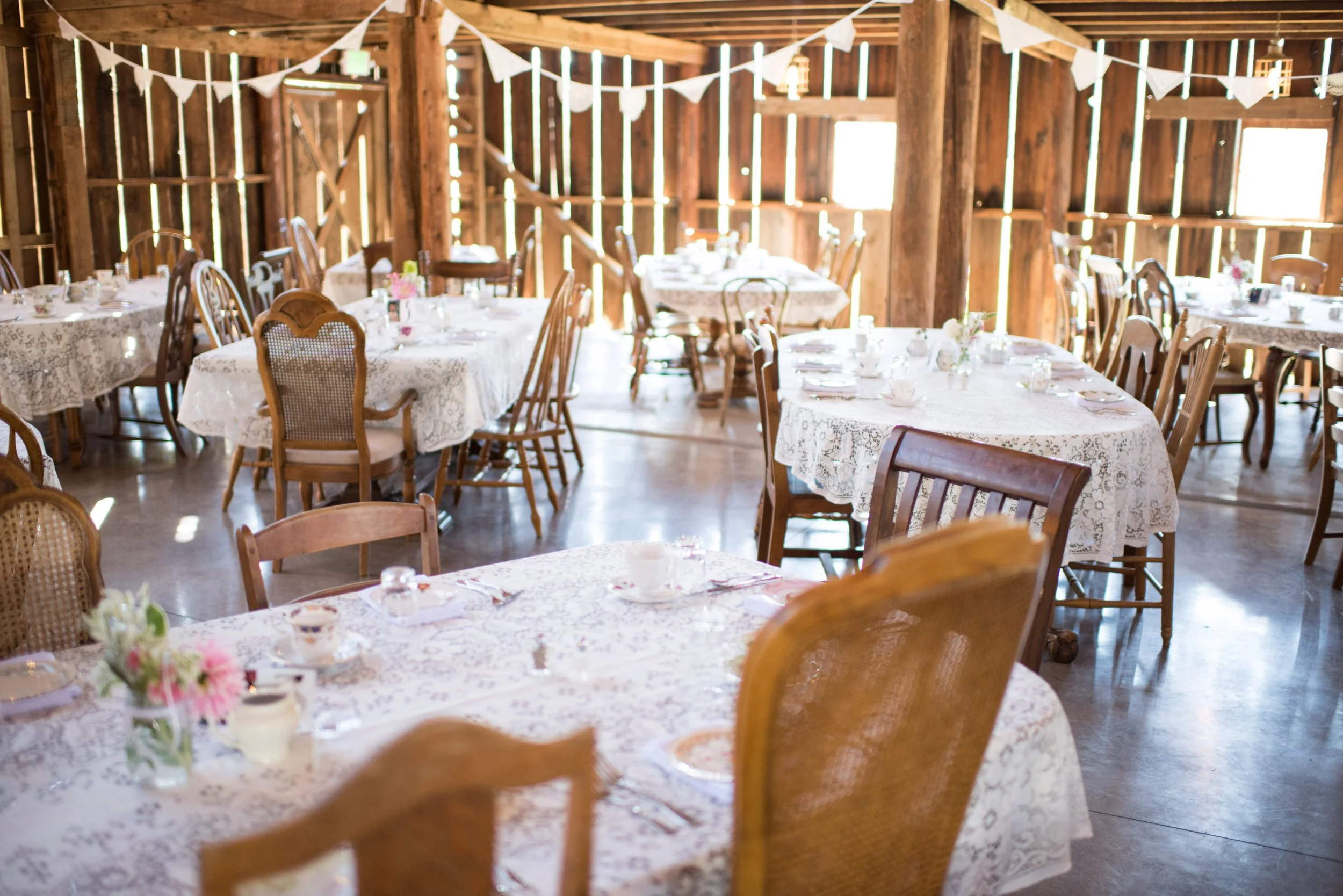 A rustic barn interior decorated for a celebration, with round and rectangular tables covered with lace tablecloths, set with plates, glasses, and floral centerpieces, and white bunting strung across the wooden beams.