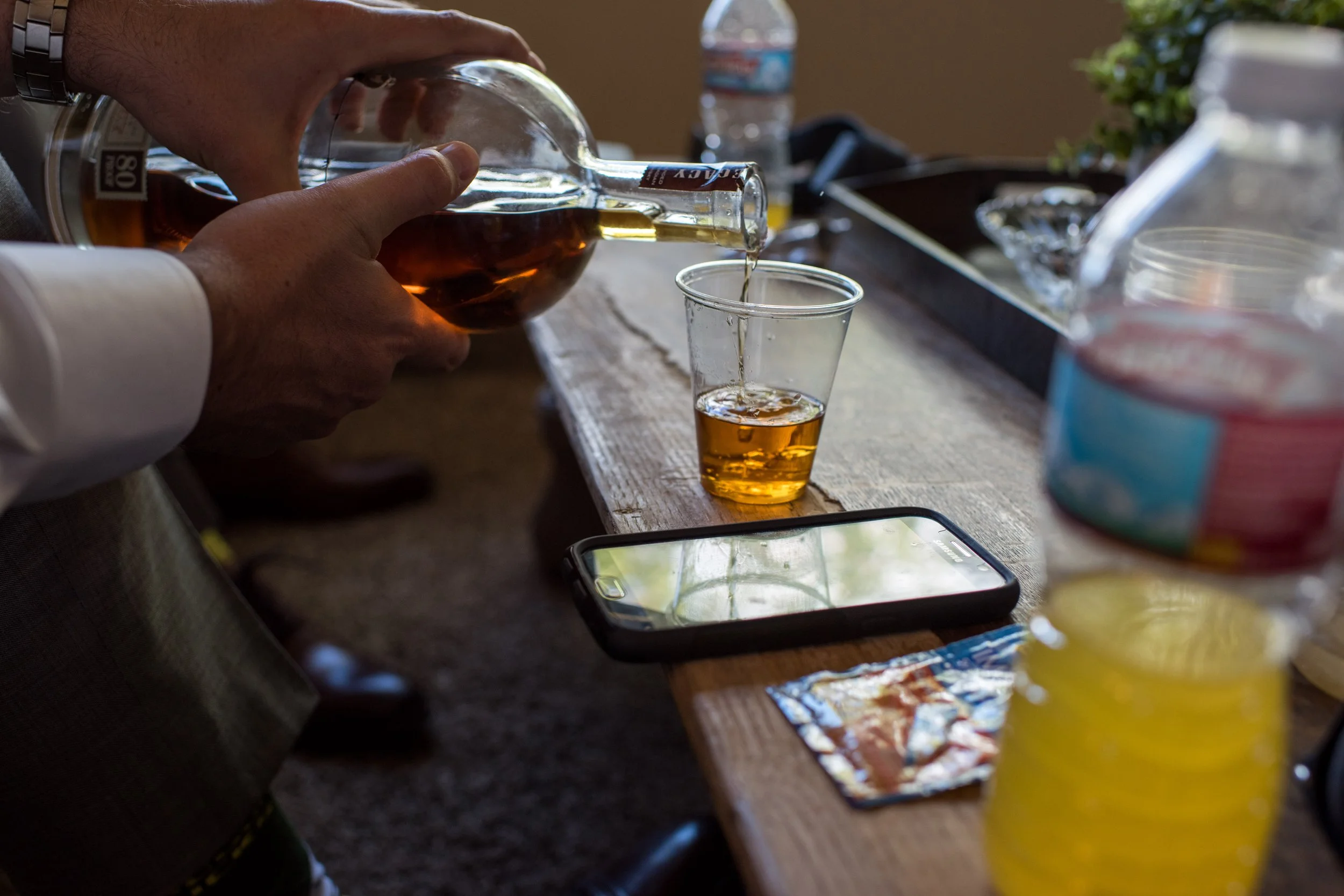 A person pouring whiskey from a glass bottle into a plastic cup on a wooden table, with various snacks and a smartphone nearby, and water bottles and a potted plant in the background.