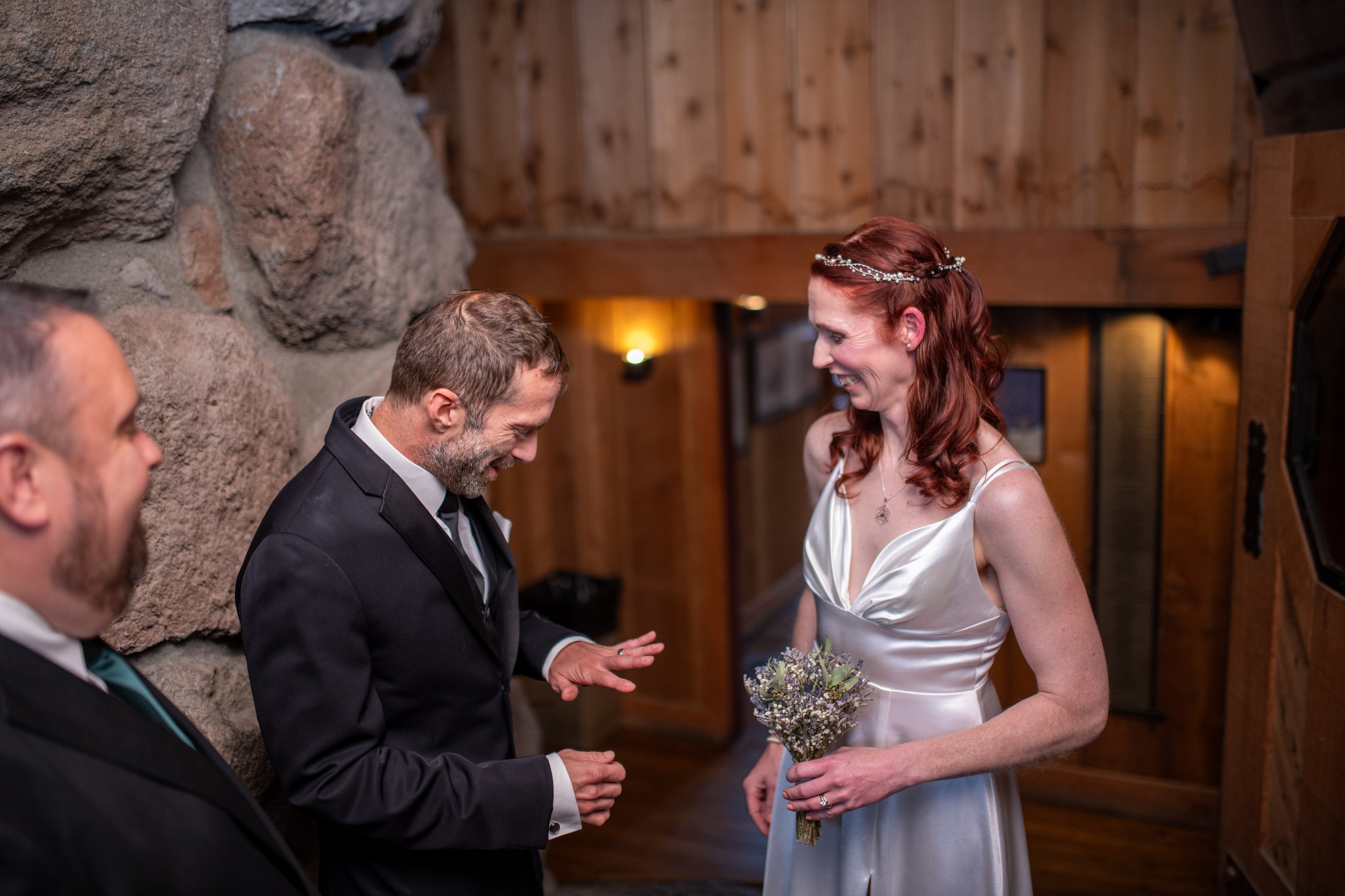 A woman in a white dress with a flower crown and long red hair smiles at a man in a dark tuxedo, who is holding her hand during a wedding ceremony inside a wooden venue with a stone wall.