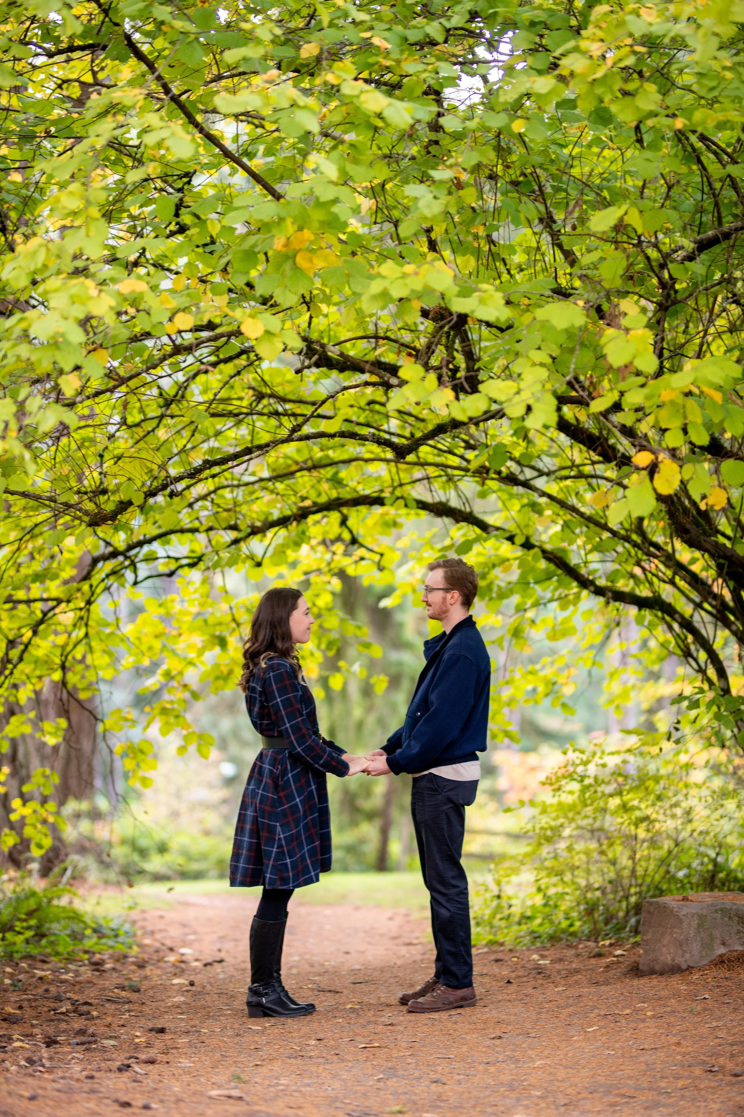 A man and a woman holding hands and facing each other under a large tree with green and yellow leaves. They are standing on a dirt path in a park.