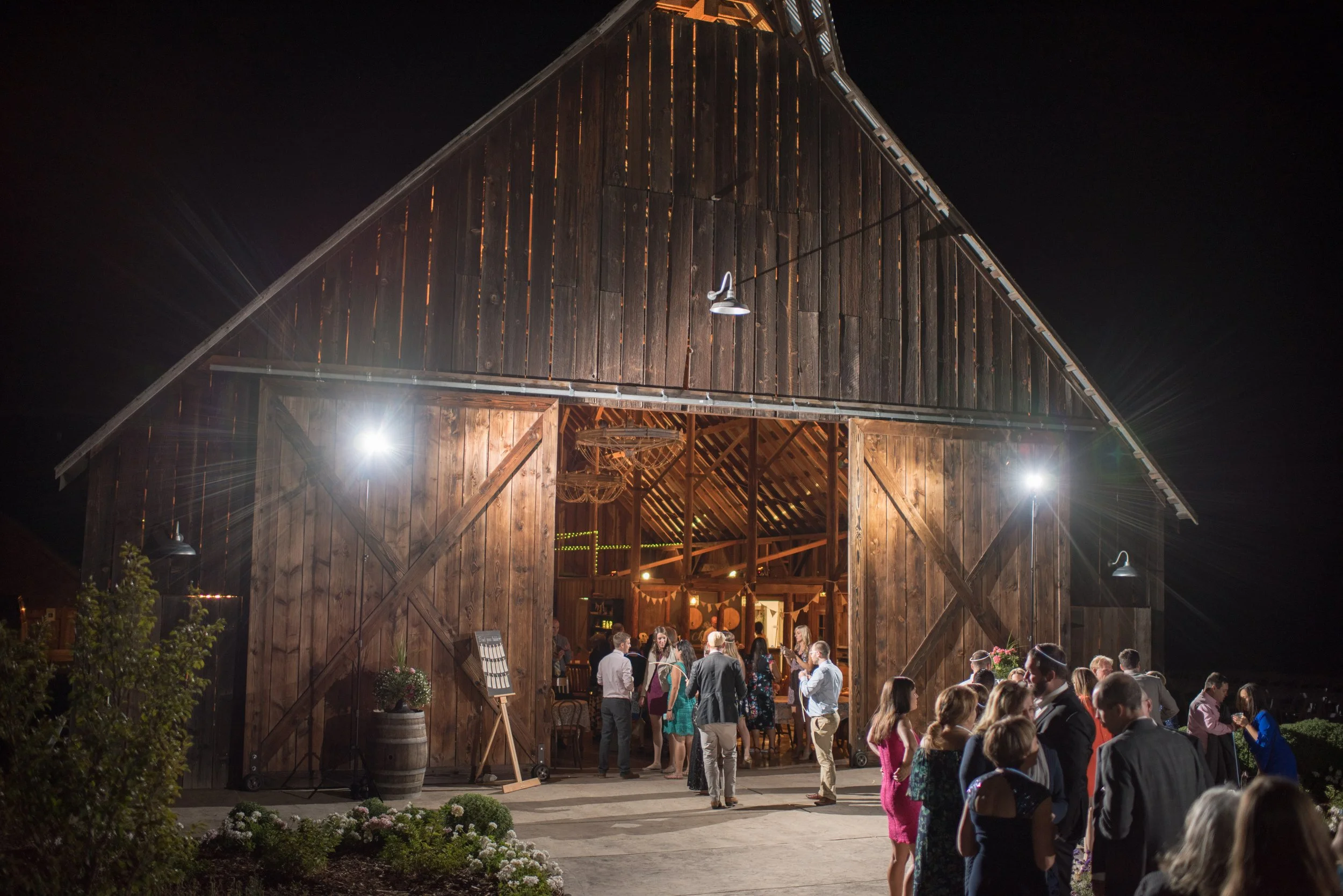 Nighttime celebration inside a rustic wooden barn with guests socializing outside and lantern lights illuminating the scene.